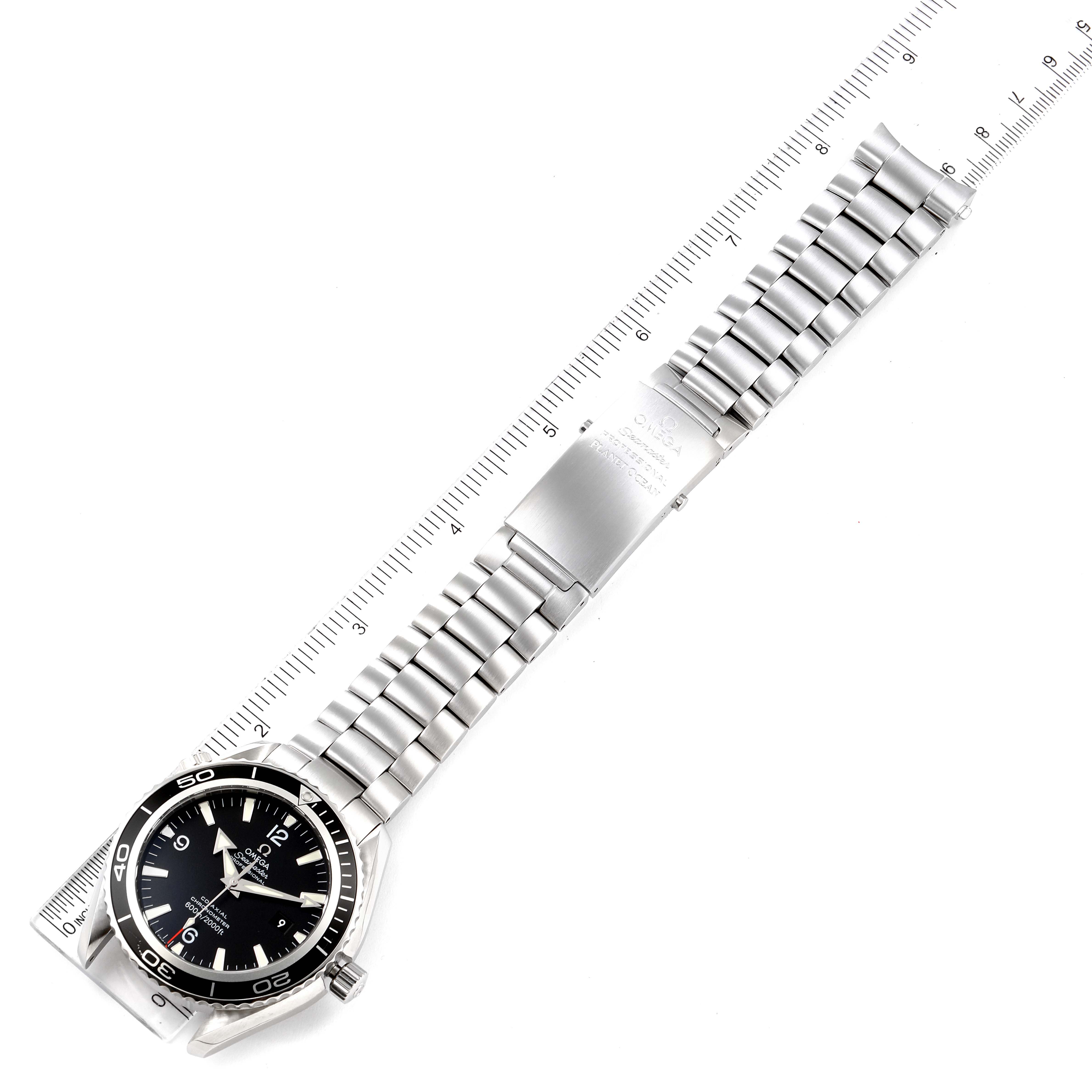 The image shows the Omega Planet Ocean 2200.50.00 Men's Stainless Steel Black Dial 2200.50.00 Men's Stainless Steel Black Dial watch laid flat against a ruler, highlighting its stainless steel bracelet and clasp.