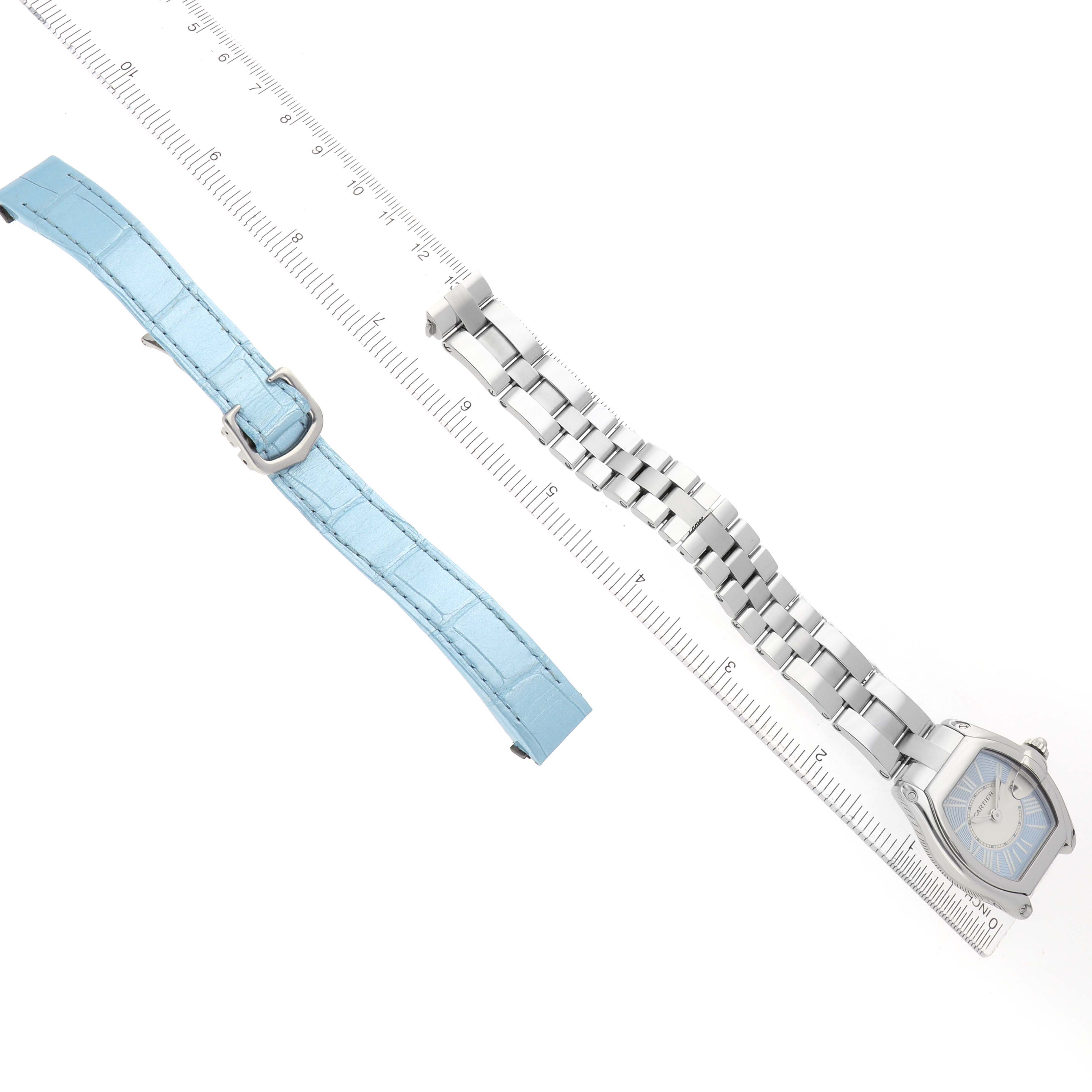 The image shows a Cartier Roadster W62053V3 Women's Stainless Steel Blue Dial watch with a metal bracelet and an additional blue leather strap, both aligned with a ruler.