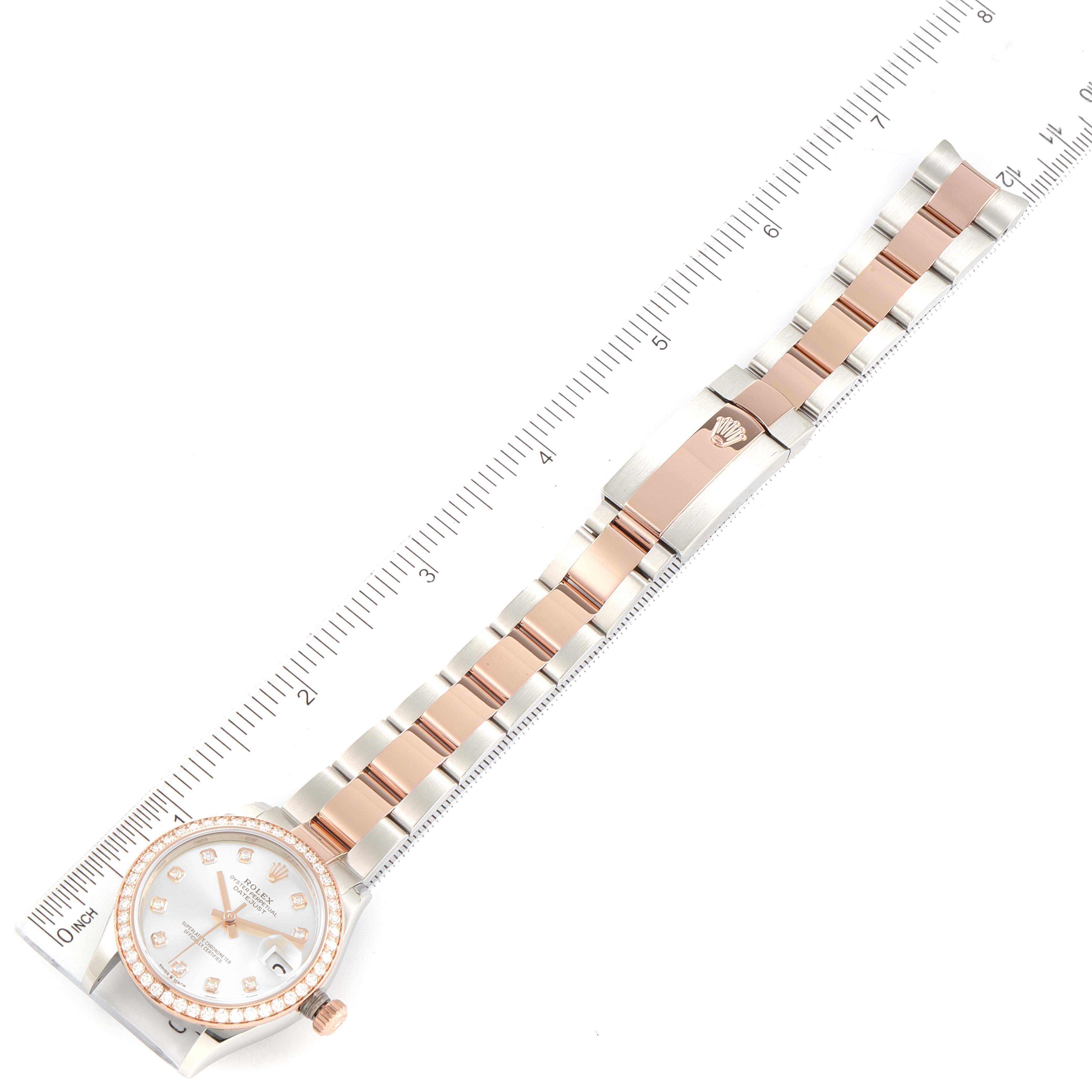 The image features a top-down view of a Rolex Mid-Size 278381 Women
s Steel and Gold (two tone) Silver Dial 278381 Women
s Steel and Gold (two tone) Silver Dial watch, displaying its face, bezel, band, and clasp against a ruler.