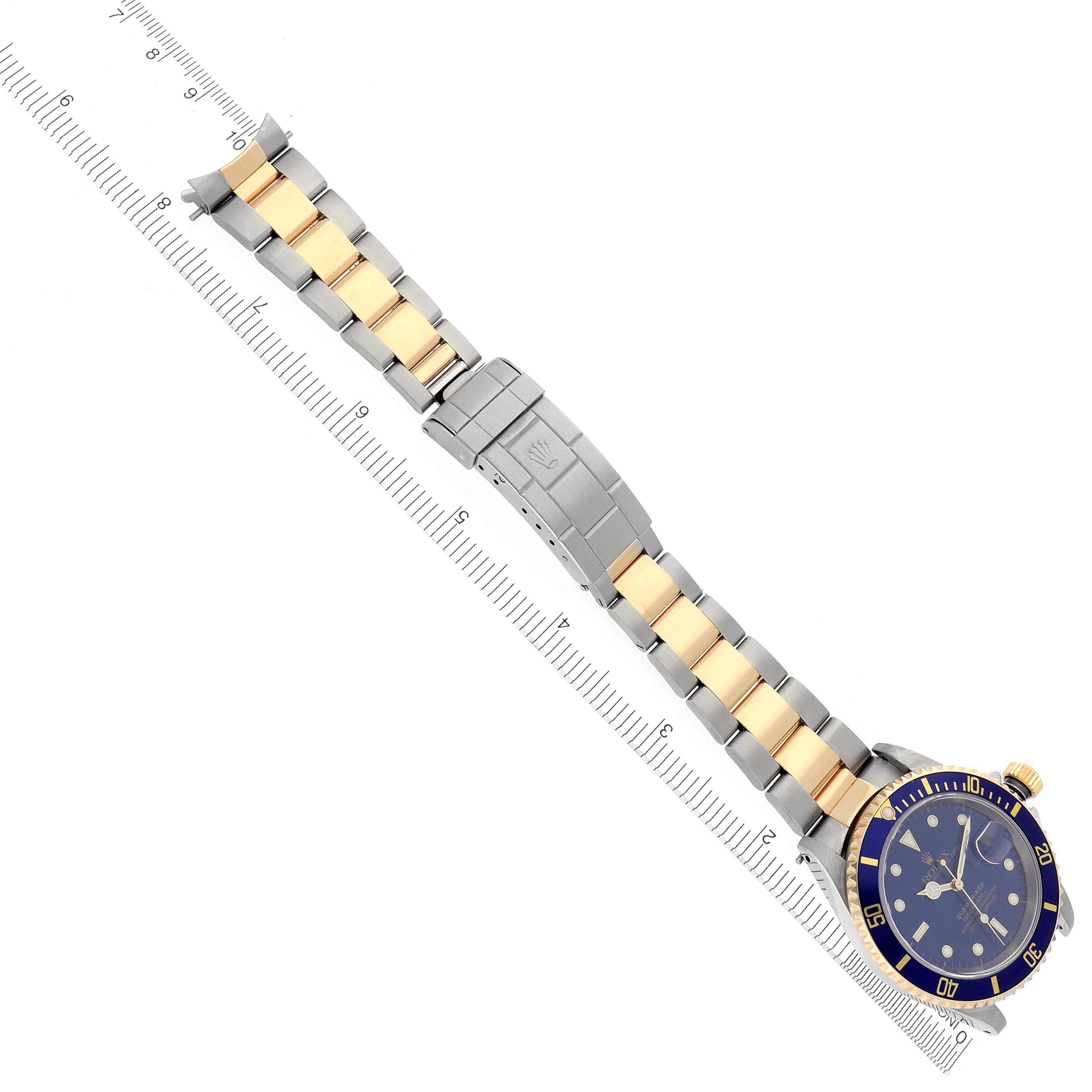 The Rolex Submariner 16613 Men's Steel and Gold (two tone) Blue Dial 16613 Men's Steel and Gold (two tone) Blue Dial watch is shown from a top-down angle, highlighting its blue and gold bezel, dial, and two-tone bracelet alongside a ruler.