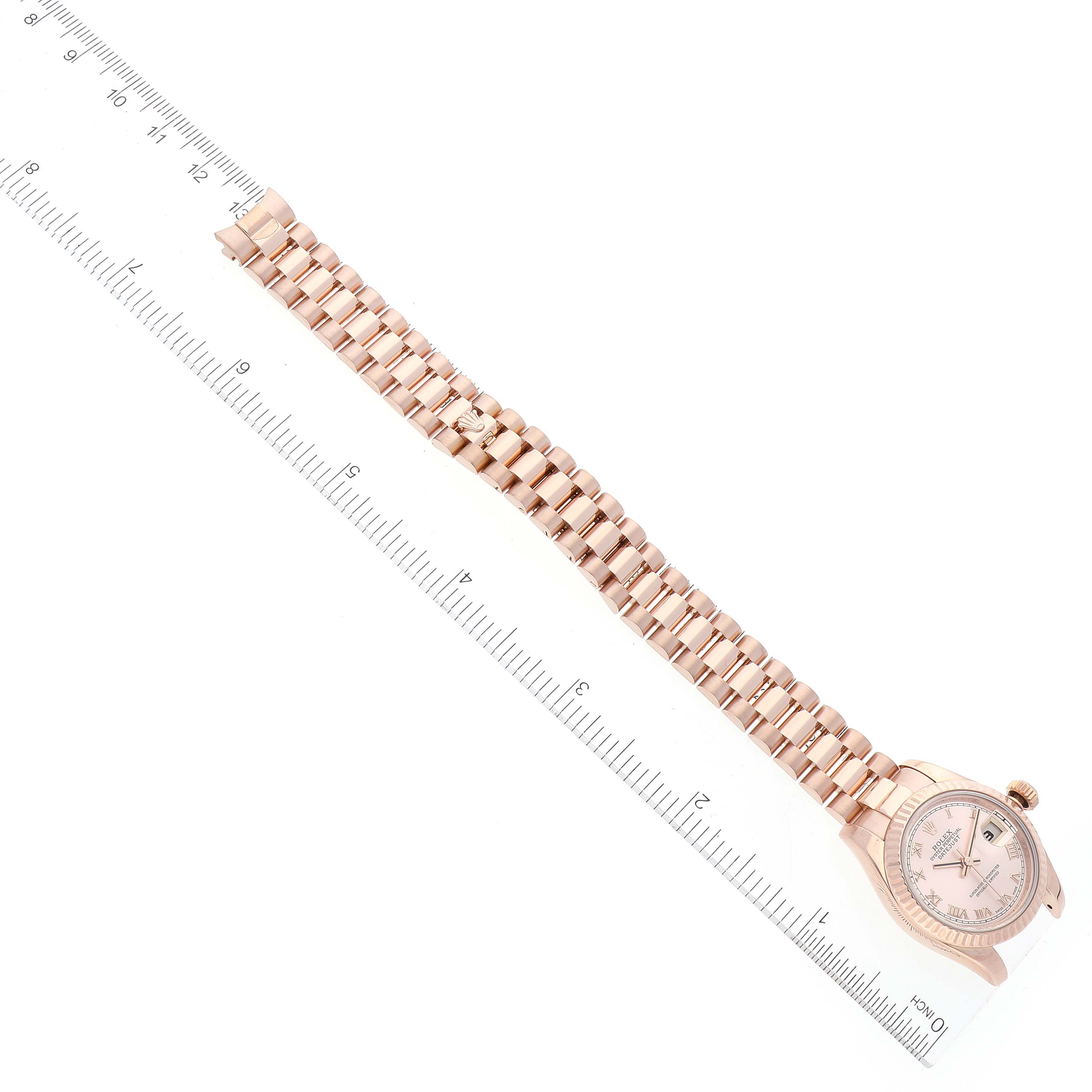 The image shows a laid-flat Rolex President 179175 Women's Rose Gold Pink Dial 179175 Women's Rose Gold Pink Dial model watch, angled from above, featuring its face and full metal bracelet against a ruler.
