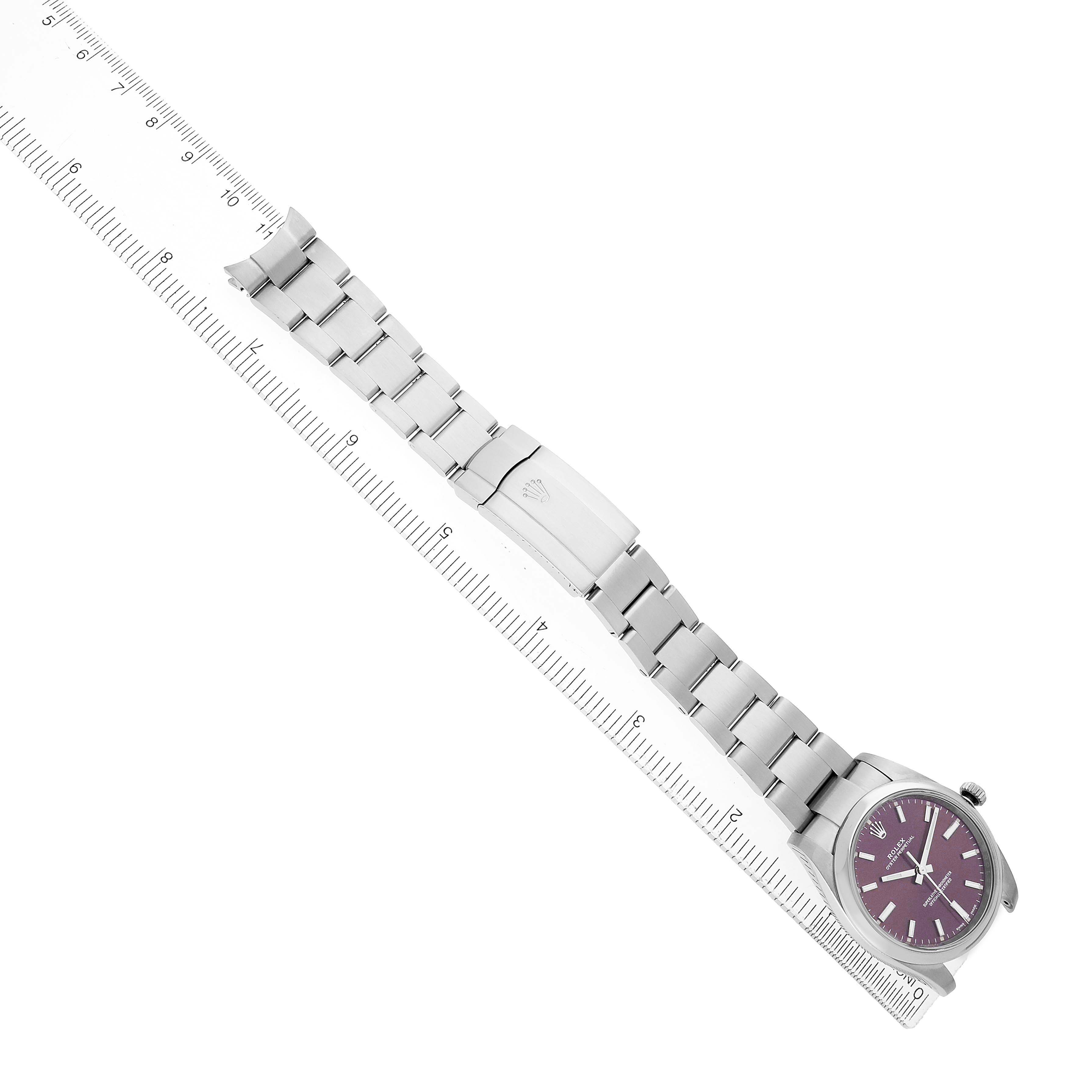 The Rolex Oyster Perpetual 114200 Men's Stainless Steel Purple Dial watch is shown at an angle from above, featuring the face, bracelet, and clasp on a ruler.