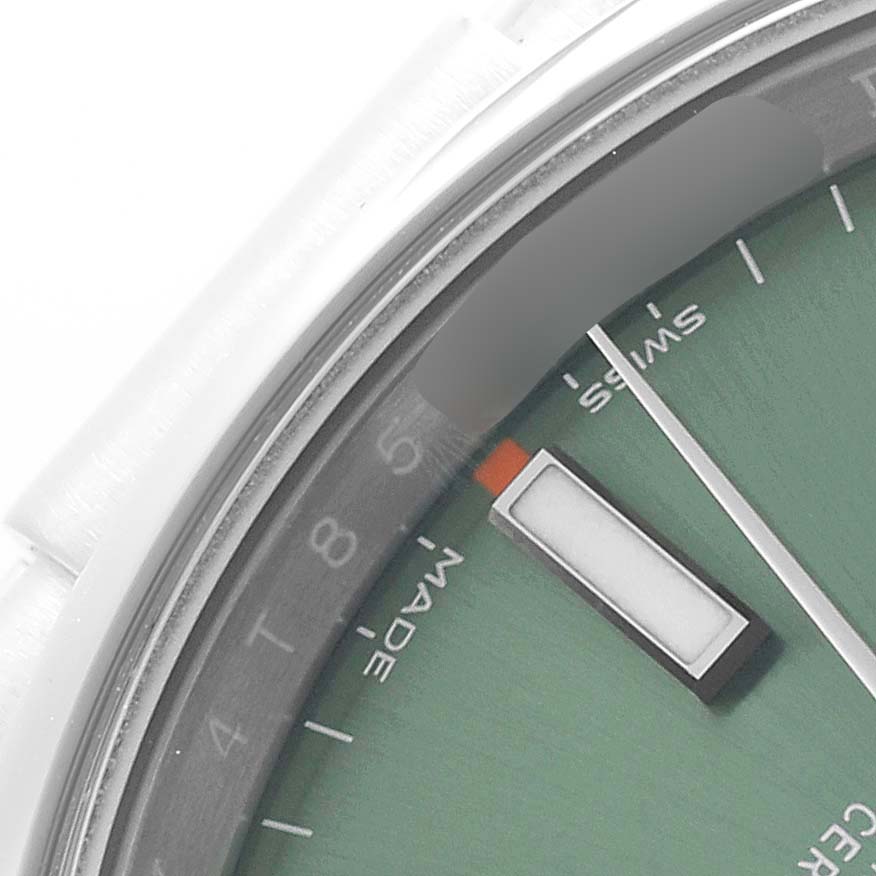 The image shows a close-up of the dial and minute hand of a Rolex Oyster Perpetual 114200 Men's Stainless Steel Green Dial watch, focusing on indices and text.