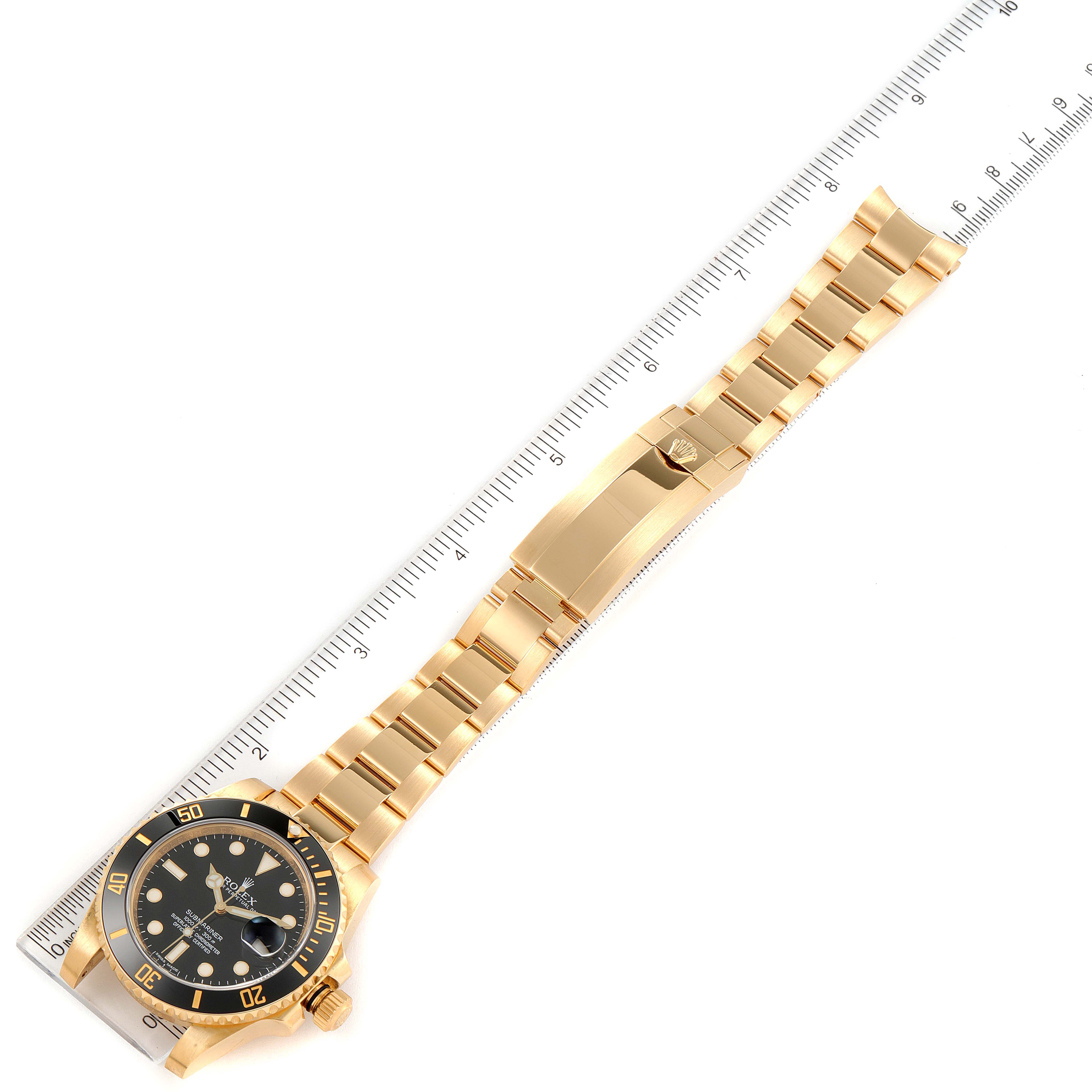 The image shows a Rolex Submariner 116618 Men
s Yellow Gold Black Dial 116618 Men
s Yellow Gold Black Dial watch lying flat with its bracelet fully extended alongside a ruler.