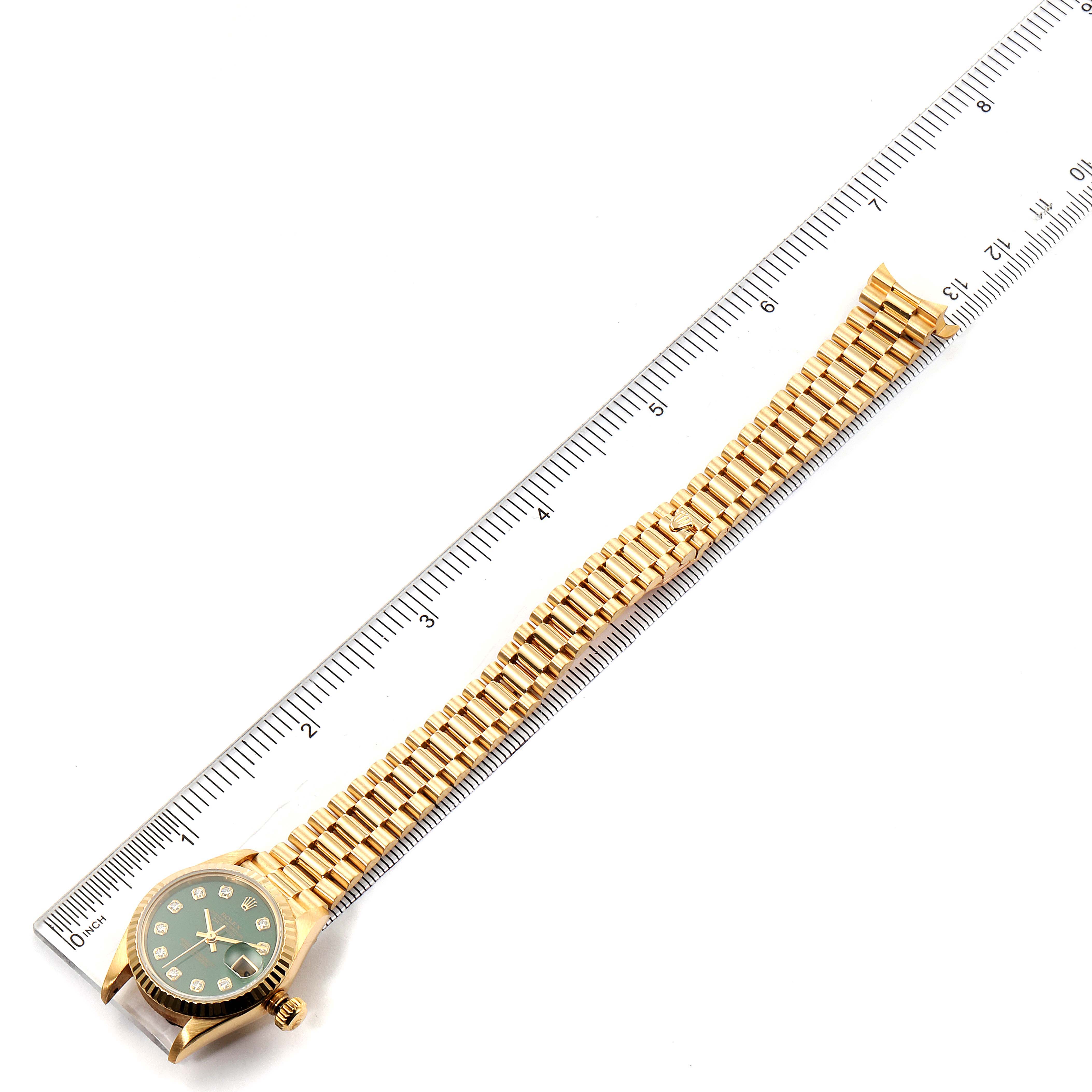 The image shows a President model Rolex watch laid flat next to a ruler, displaying its face and full gold bracelet.