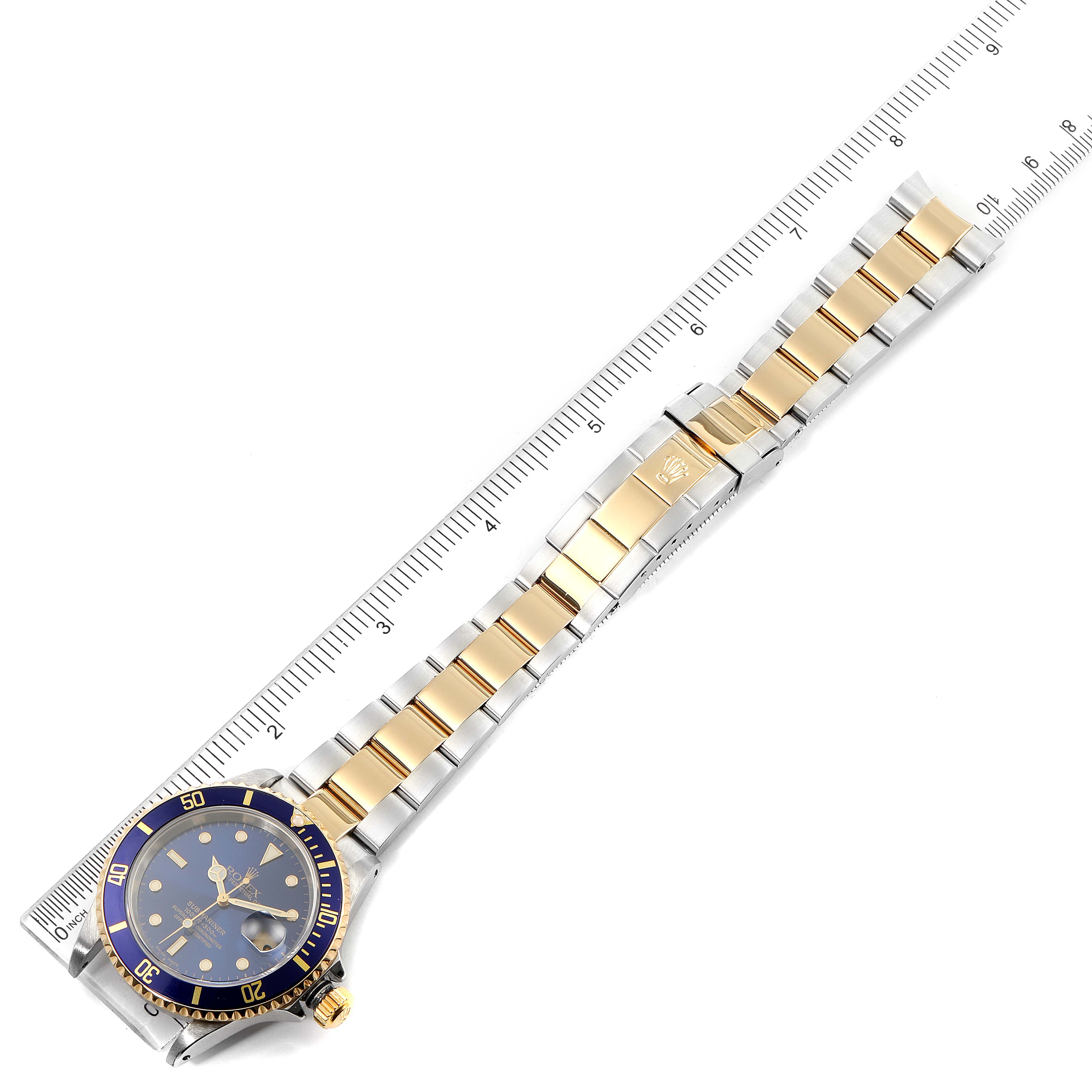 The image shows a Rolex Submariner 16613 Men's Steel and Gold (two tone) Blue Dial 16613 Men's Steel and Gold (two tone) Blue Dial watch laid flat, highlighting its band, bezel, and face, next to a ruler for scale.