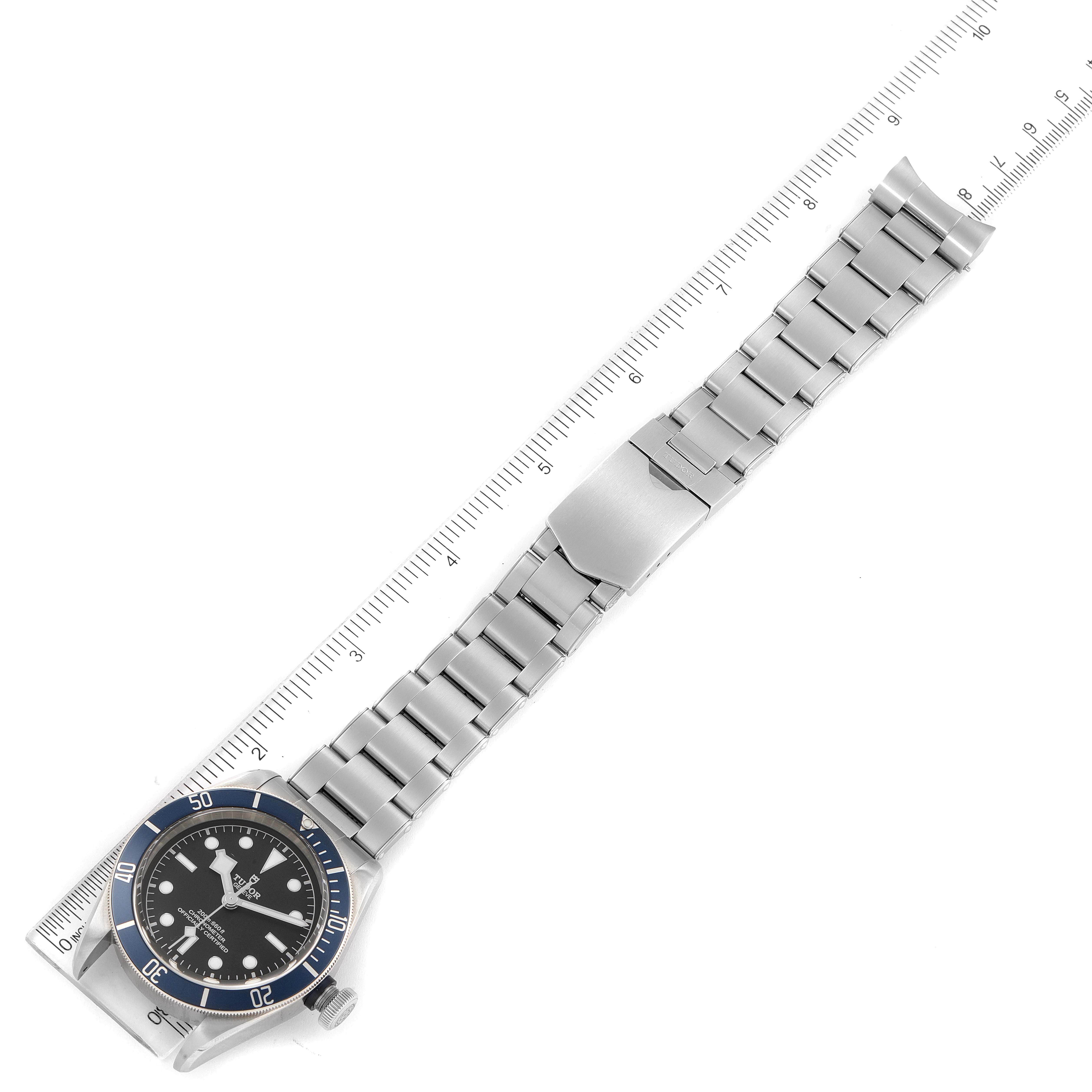 The Tudor Heritage Black Bay 79230B Men's Stainless Steel Black Dial 79230B Men's Stainless Steel Black Dial watch is shown at a top-down angle, featuring the watch face and metal bracelet with a ruler for scale.