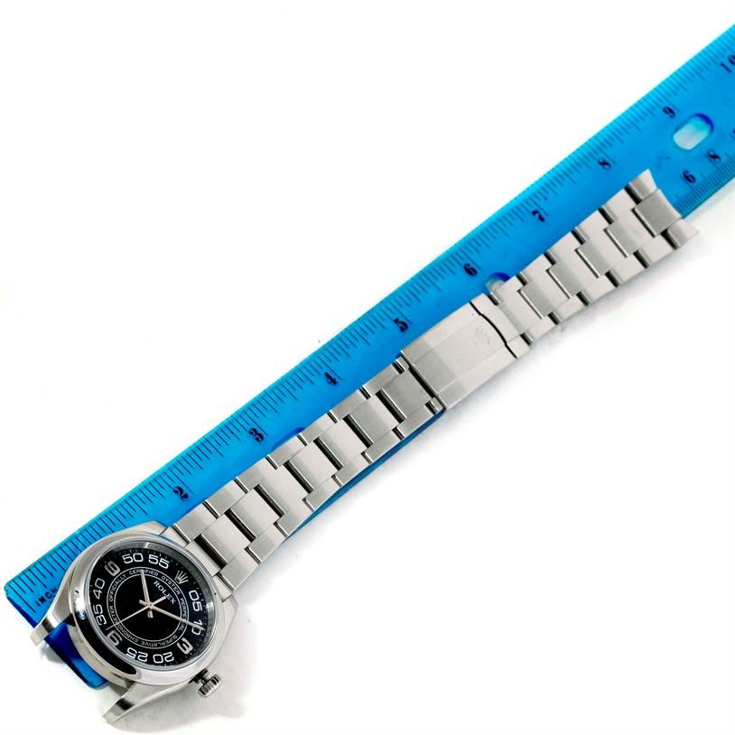 The image shows a top-down view of a Rolex Oyster Perpetual 116000 Men's Black Dial 116000 Men's Black Dial watch, displaying the face, casing, and bracelet alongside a blue ruler.