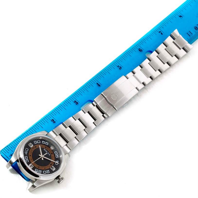 This image shows a Rolex Oyster Perpetual 116000 Men
s Stainless Steel Black Dial 116000 Men
s Stainless Steel Black Dial watch laid flat next to a blue ruler, highlighting the watch face, bracelet, and clasp.