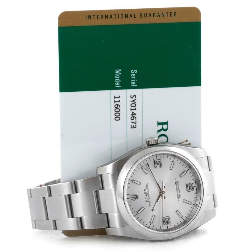 The image shows a Rolex Oyster Perpetual 116000 Men's Stainless Steel Silver Dial 116000 Men's Stainless Steel Silver Dial watch lying down with its bracelet visible and an international guarantee card in the background.