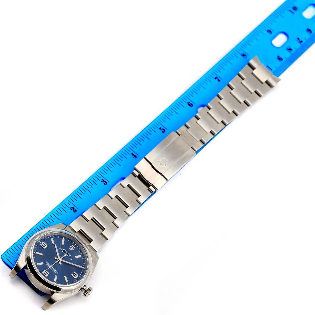 This image shows the Rolex Oyster Perpetual 116000 Men
s Stainless Steel Blue Dial 116000 Men
s Stainless Steel Blue Dial watch in a flat lay, alongside a blue ruler for scale, highlighting its face and bracelet.
