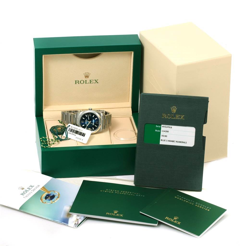 The Rolex Oyster Perpetual 114200 Men's Stainless Steel Blue Dial 114200 Men's Stainless Steel Blue Dial watch is shown in its box along with manuals, warranty card, and accessories at a front angle.