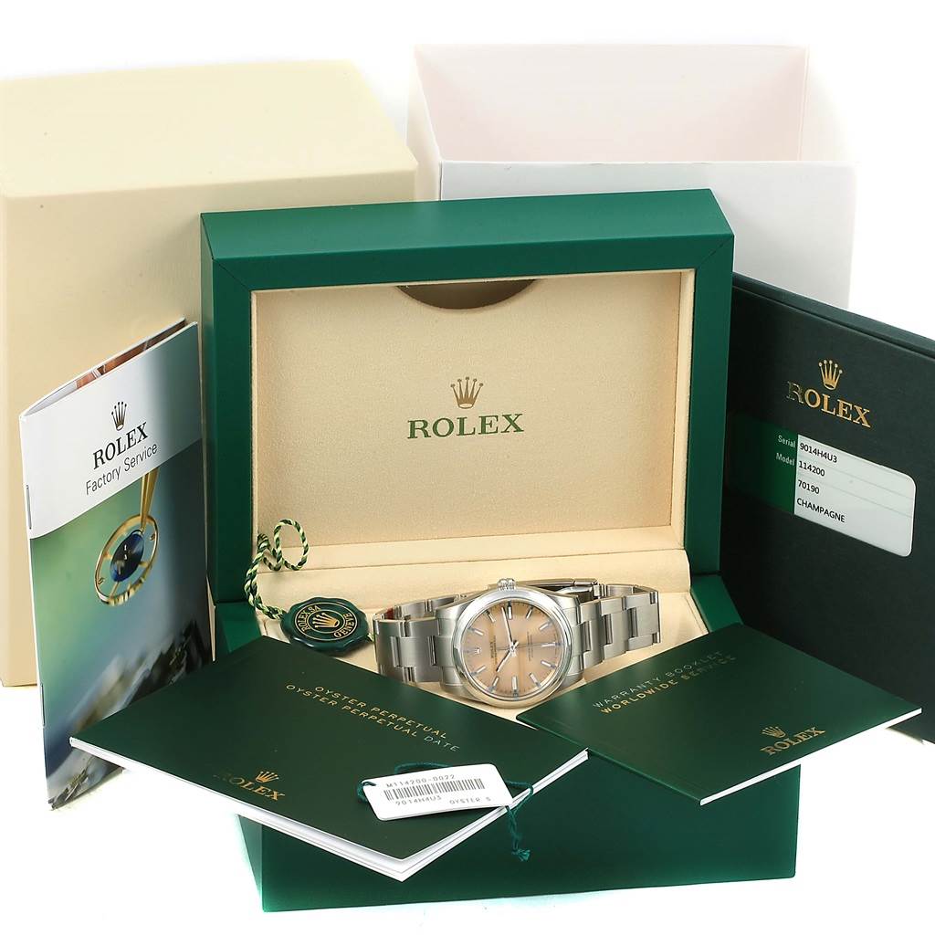 This image shows a Rolex Oyster Perpetual 114200 Men's Stainless Steel Champagne Dial 114200 Men's Stainless Steel Champagne Dial watch in its box with accompanying booklets and tags, taken from a front angle.