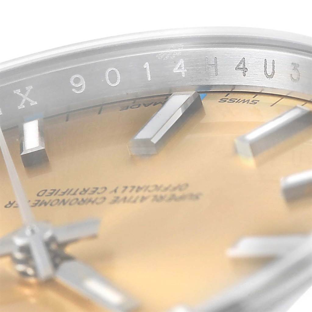 This close-up angle of the Rolex Oyster Perpetual 114200 Men's Stainless Steel Champagne Dial 114200 Men's Stainless Steel Champagne Dial shows part of the dial, index markers, and part of the bezel with a serial number.