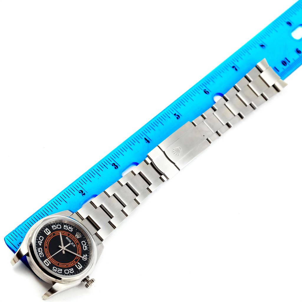 The image shows a top-down view of a Rolex Oyster Perpetual 116000 Men
s Stainless Steel Black Dial 116000 Men
s Stainless Steel Black Dial watch and its stainless steel bracelet next to a blue ruler.