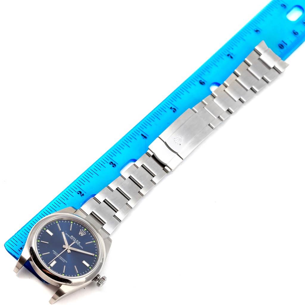 The image shows a full view of the Rolex Oyster Perpetual 114300 Men's Stainless Steel Blue Dial 114300 Men's Stainless Steel Blue Dial watch, including its blue dial, stainless steel case, and bracelet, next to a blue ruler.