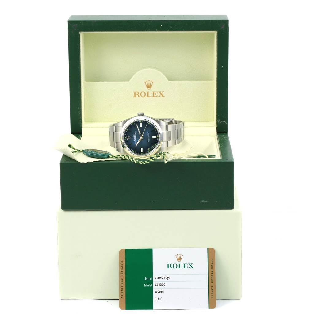 The image shows a Rolex Oyster Perpetual 114300 Men's Stainless Steel Blue Dial 114300 Men's Stainless Steel Blue Dial watch from a front angle, displayed in its open box with tags and authenticity card.