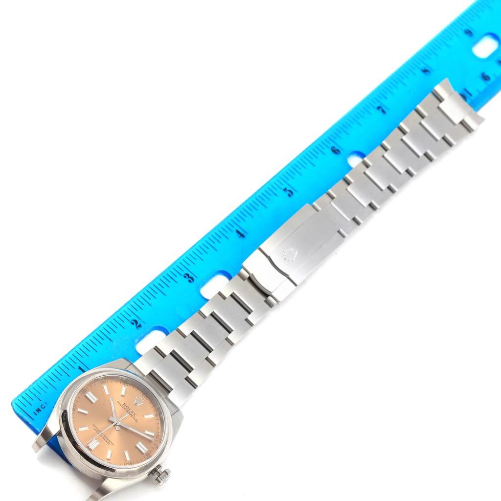 The Rolex Oyster Perpetual 116000 Men's Stainless Steel Champagne Dial 116000 Men's Stainless Steel Champagne Dial watch is shown from a top-down angle, featuring its face, bracelet, and clasp, alongside a blue ruler for scale.