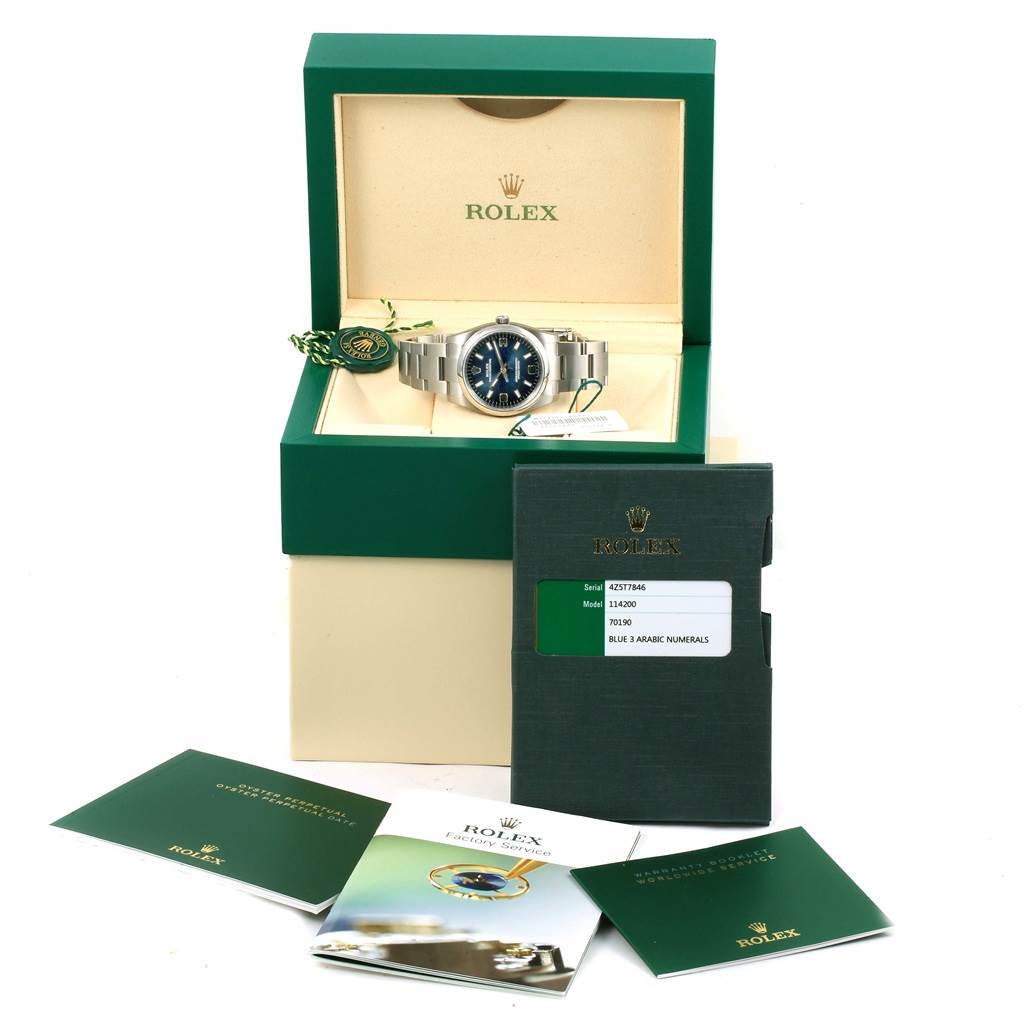 The Rolex Oyster Perpetual 114200 Men's Stainless Steel Blue Dial 114200 Men's Stainless Steel Blue Dial watch is displayed in its open green box with accompanying booklets and warranty card visible in front.