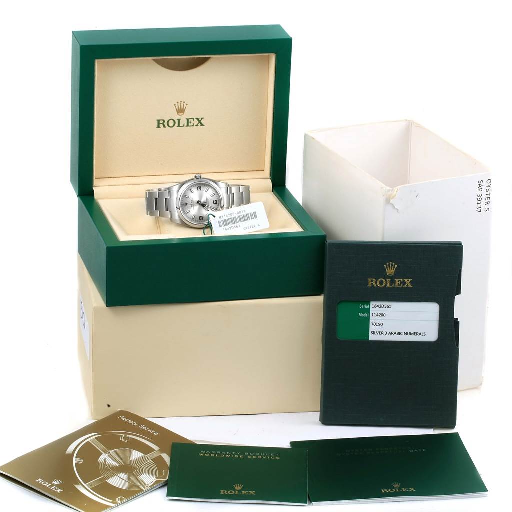 The image shows a Rolex Oyster Perpetual 114200 Stainless Steel Silver Dial 114200 Stainless Steel Silver Dial watch in its open box, with warranty booklet and tags visible from a front angle.