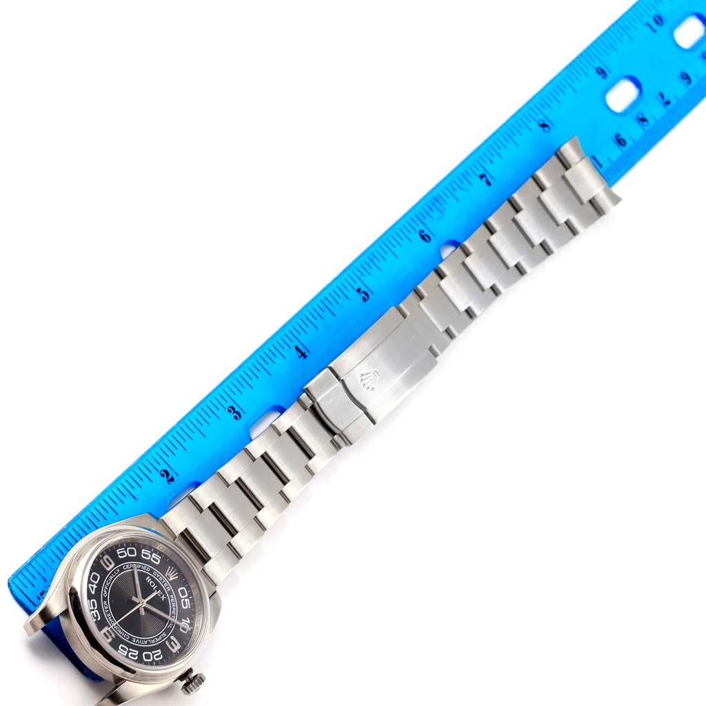 The image shows a top-down view of a Rolex Oyster Perpetual 116000 Men's Stainless Steel Black Dial 116000 Men's Stainless Steel Black Dial watch displaying the face, bracelet, and clasp alongside a ruler.