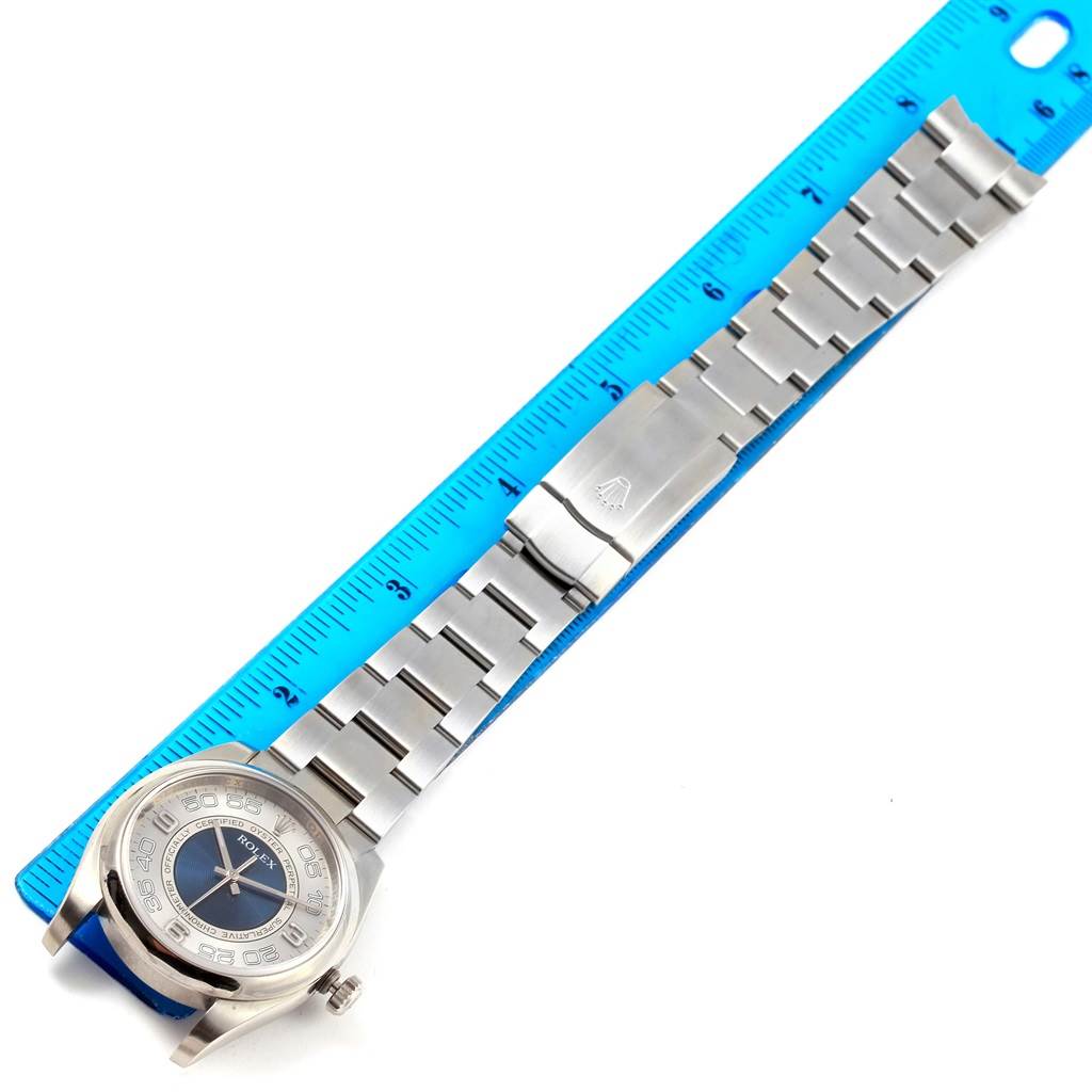 The image displays an overhead view of a Rolex Oyster Perpetual 116000 Men's Stainless Steel Silver Dial 116000 Men's Stainless Steel Silver Dial watch, showcasing its face, bracelet, and clasp alongside a blue measuring ruler.