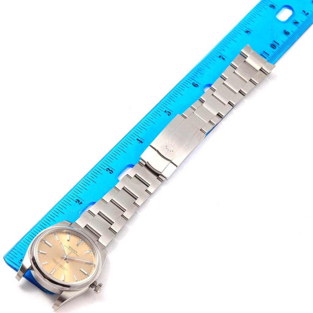The Rolex Oyster Perpetual 114200 Stainless Steel Champagne Dial 114200 Stainless Steel Champagne Dial watch is shown from a top-down angle, displaying the face, bracelet, and clasp, next to a ruler for scale.