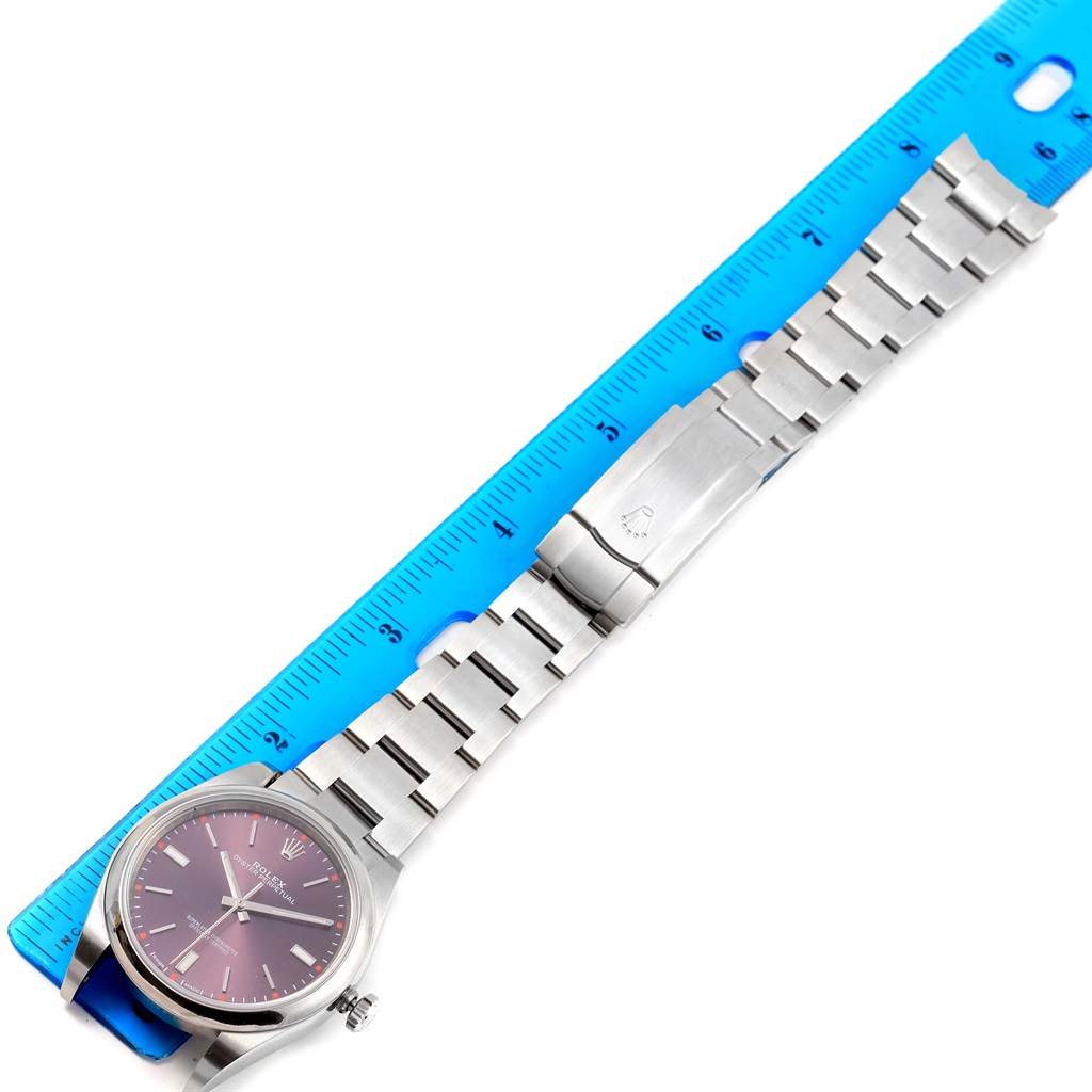 The image shows a top-down view of a Rolex Oyster Perpetual 114300 Stainless Steel Burgundy Dial 114300 Stainless Steel Burgundy Dial watch and its stainless steel bracelet next to a blue ruler.