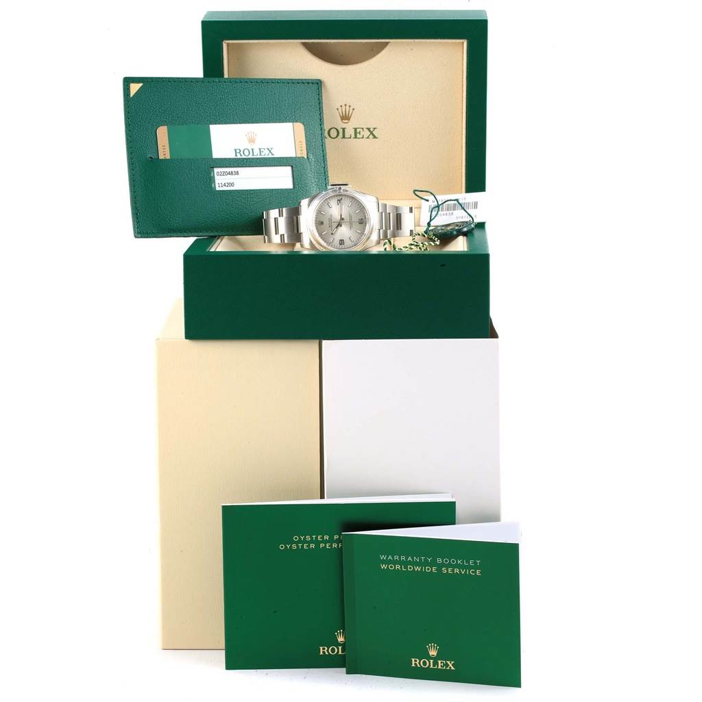 The image shows a Rolex Oyster Perpetual 114200 Stainless Steel Silver Dial 114200 Stainless Steel Silver Dial watch with its box, warranty card, and booklets viewed from the front.