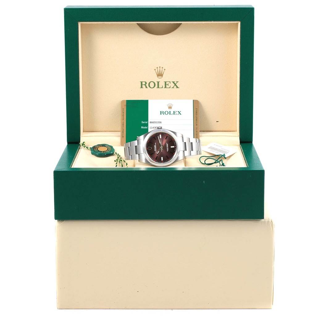 The image shows a Rolex Oyster Perpetual 114300 Stainless Steel Burgundy Dial 114300 Stainless Steel Burgundy Dial watch at a front angle within its box, with accompanying warranty card and accessories.