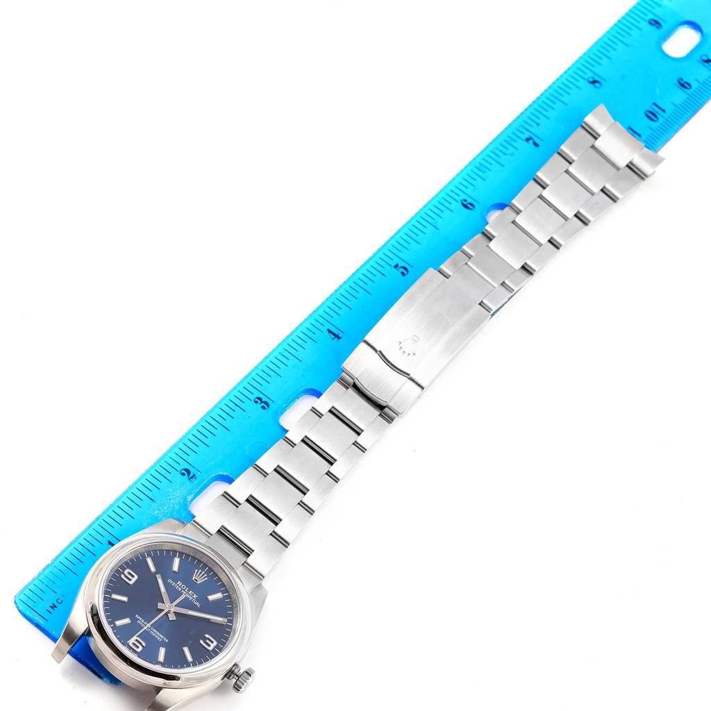 The image shows an overhead view of a Rolex Oyster Perpetual 116000 Men's Stainless Steel Blue Dial 116000 Men's Stainless Steel Blue Dial watch with a stainless steel band placed next to a blue ruler.