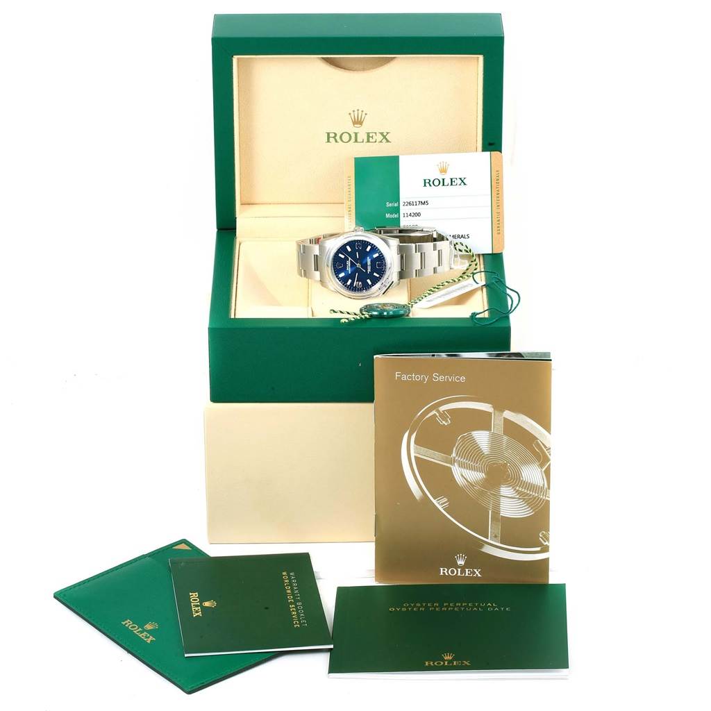The image shows a Rolex Oyster Perpetual 114200 Men's Stainless Steel Blue Dial 114200 Men's Stainless Steel Blue Dial watch in its box, displayed with warranty card, manuals, and factory service booklet.