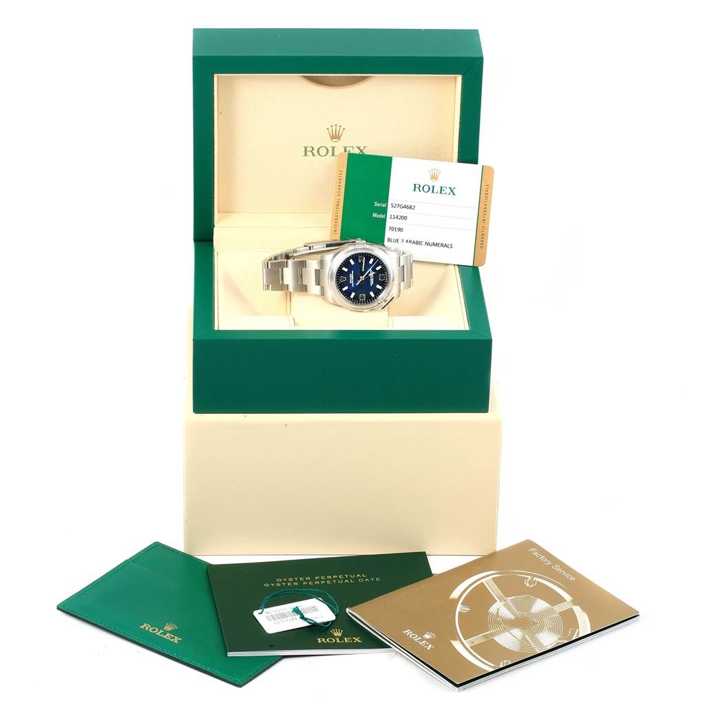 The image shows the Rolex Oyster Perpetual 114200 Men's Stainless Steel Blue Dial 114200 Men's Stainless Steel Blue Dial watch in a display box, with the warranty card and instructional booklets placed around it.