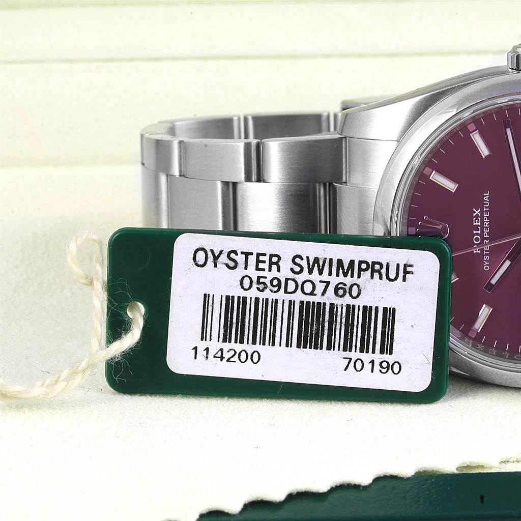 The image shows a partial view of the Rolex Oyster Perpetual 114200 Stainless Steel Burgundy Dial 114200 Stainless Steel Burgundy Dial watch, focusing on the strap, case, and a tag with model details.