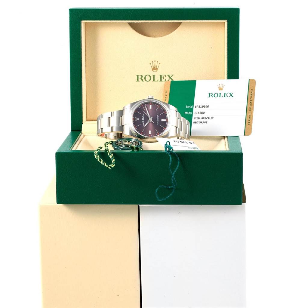 The Rolex Oyster Perpetual 114300 Stainless Steel Burgundy Dial 114300 Stainless Steel Burgundy Dial watch is shown from the front in its open green box, along with its certification card and accessories.