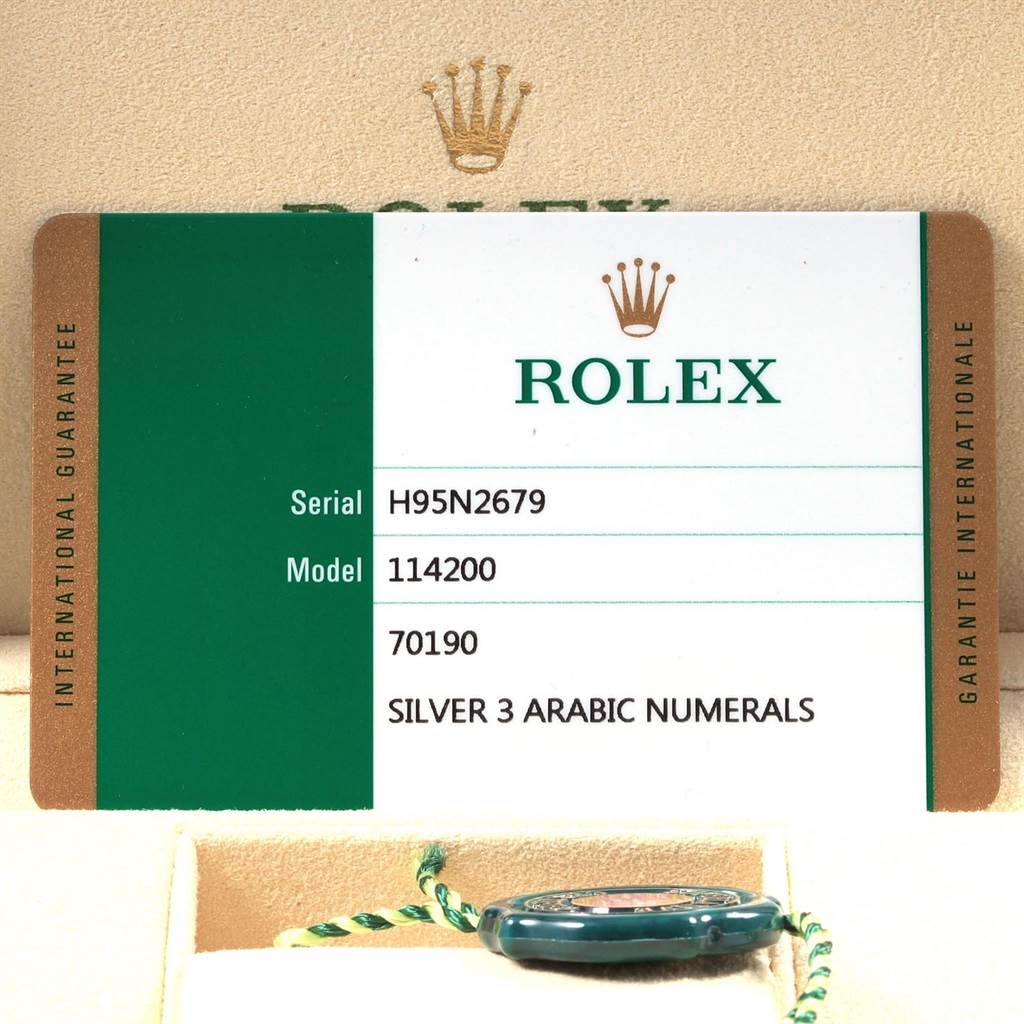 The image shows the warranty card for a Rolex Oyster Perpetual 114200 Men's Stainless Steel Silver Dial 114200 Men's Stainless Steel Silver Dial model 114200 and a partially visible green tag.