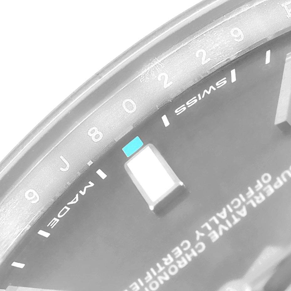 The image shows a close-up of the dial on the Rolex Oyster Perpetual 114300 Men
s Stainless Steel Grey Dial 114300 Men
s Stainless Steel Grey Dial watch, featuring part of the minute markers, the hour markers, and text on the dial.