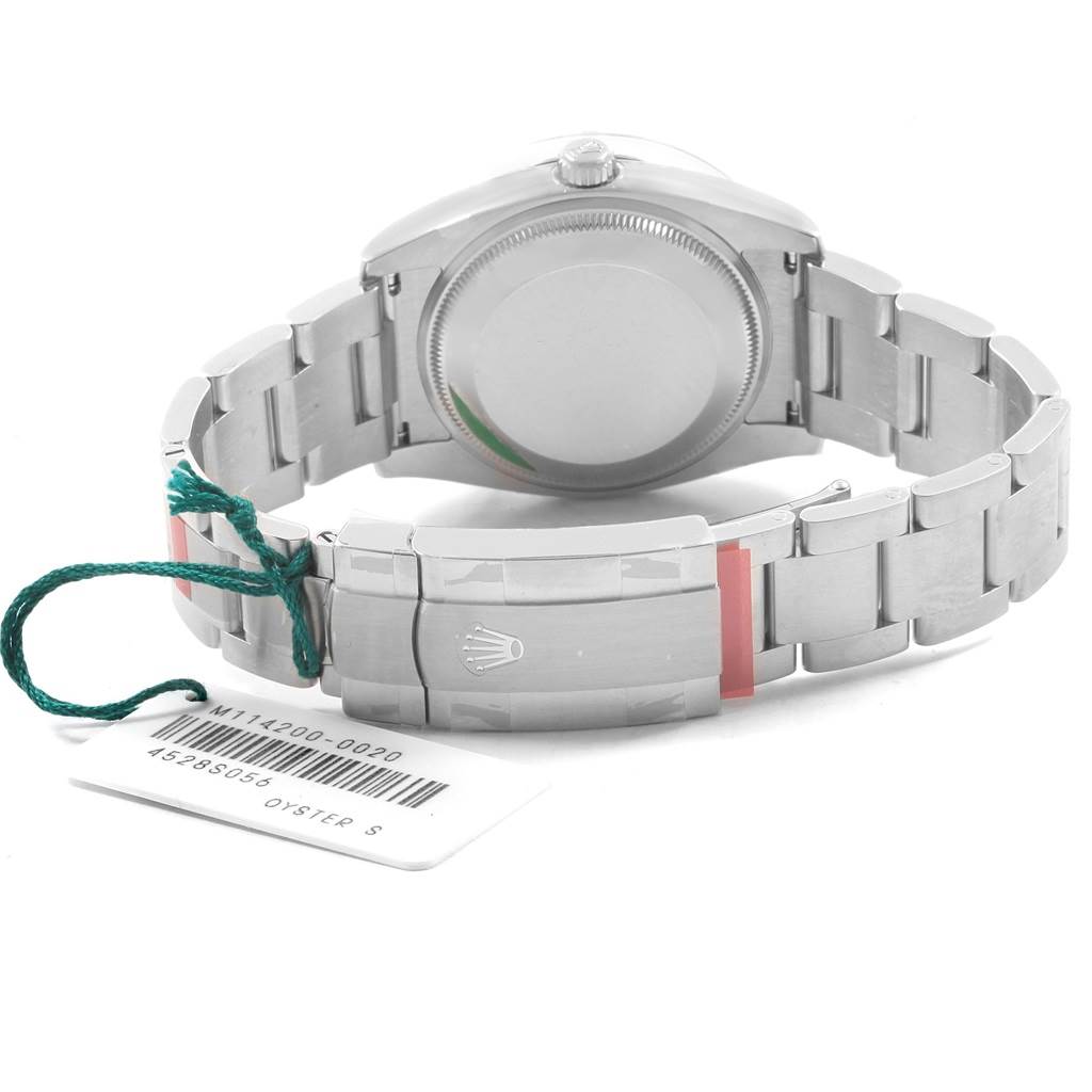 The image shows the back view of a Rolex Oyster Perpetual 114200 Men
s Stainless Steel Burgundy Dial 114200 Men
s Stainless Steel Burgundy Dial watch, focusing on the case back and the clasp with an attached tag.