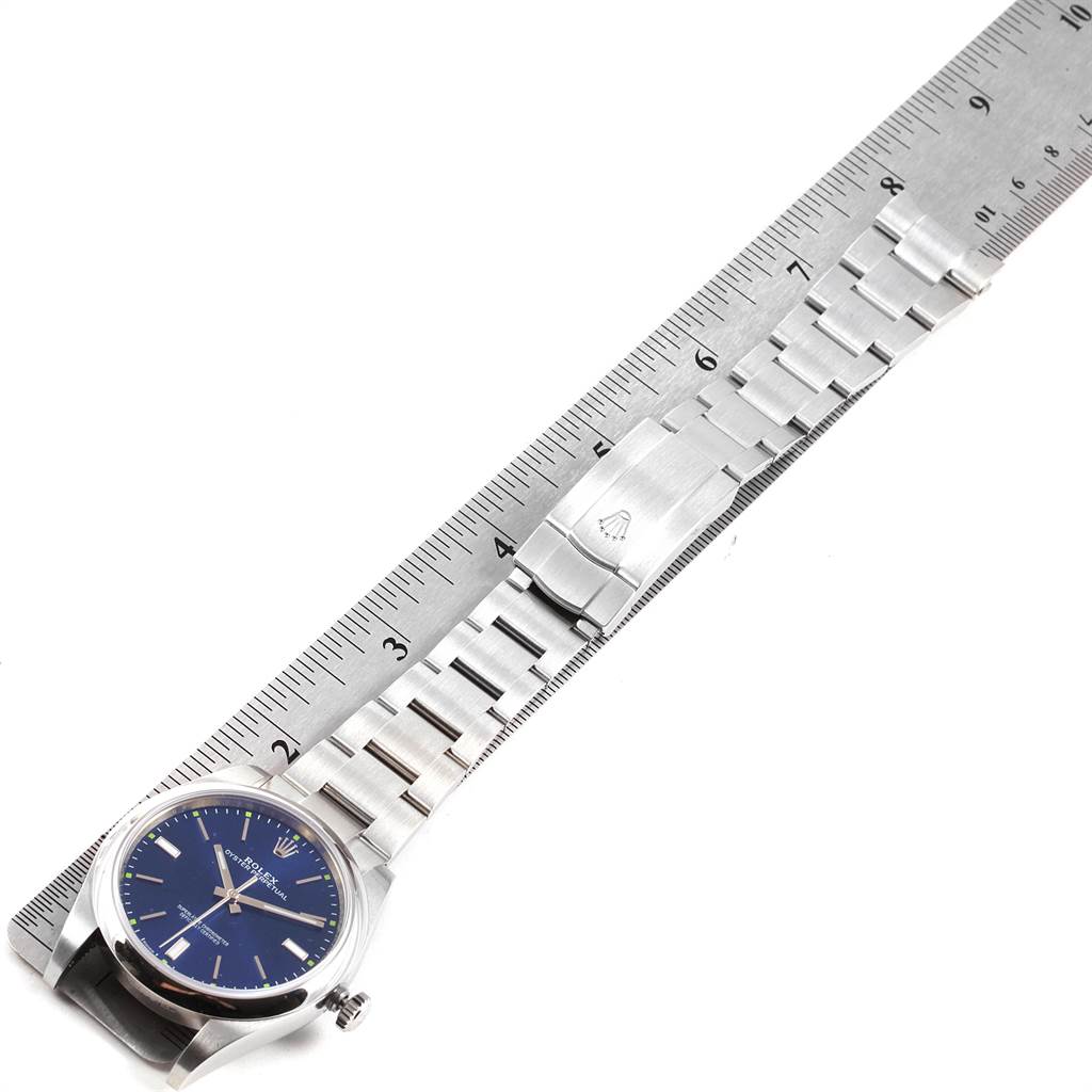The image shows a Rolex Oyster Perpetual 114300 Men
s Stainless Steel Blue Dial 114300 Men
s Stainless Steel Blue Dial watch laid flat along a ruler, showcasing the watch face and metal bracelet.
