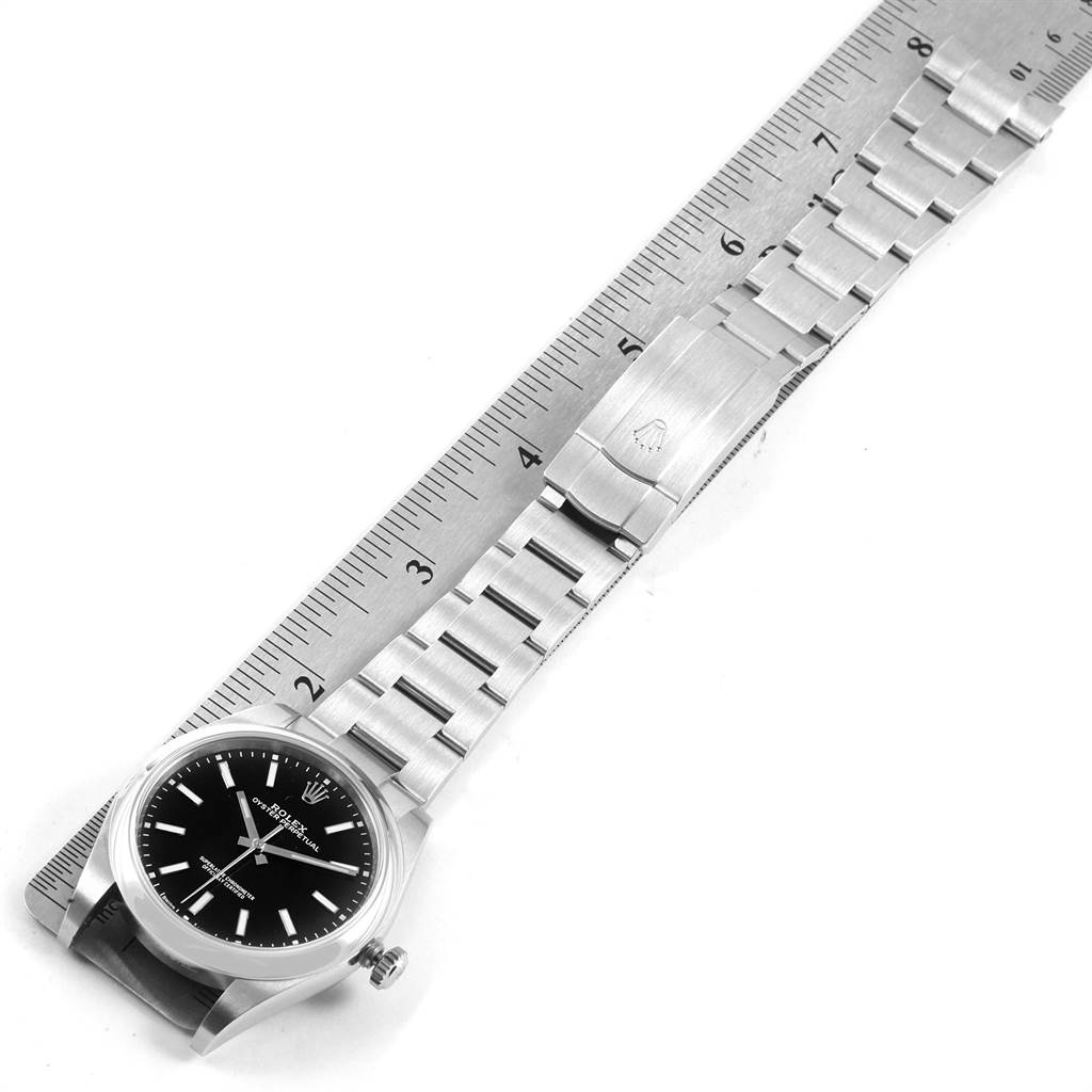 The Rolex Oyster Perpetual 114300 Men
s Stainless Steel Black Dial 114300 Men
s Stainless Steel Black Dial watch is shown from a top-down angle, displaying its face, bracelet, clasps, and part of a ruler measuring its length.