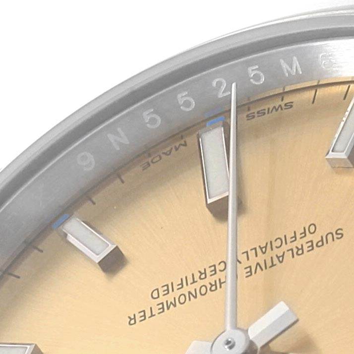 This close-up image of the Rolex Oyster Perpetual 114200 Men's Stainless Steel Champagne Dial 114200 Men's Stainless Steel Champagne Dial watch shows the dial, hour markers, and partial text at an angle.