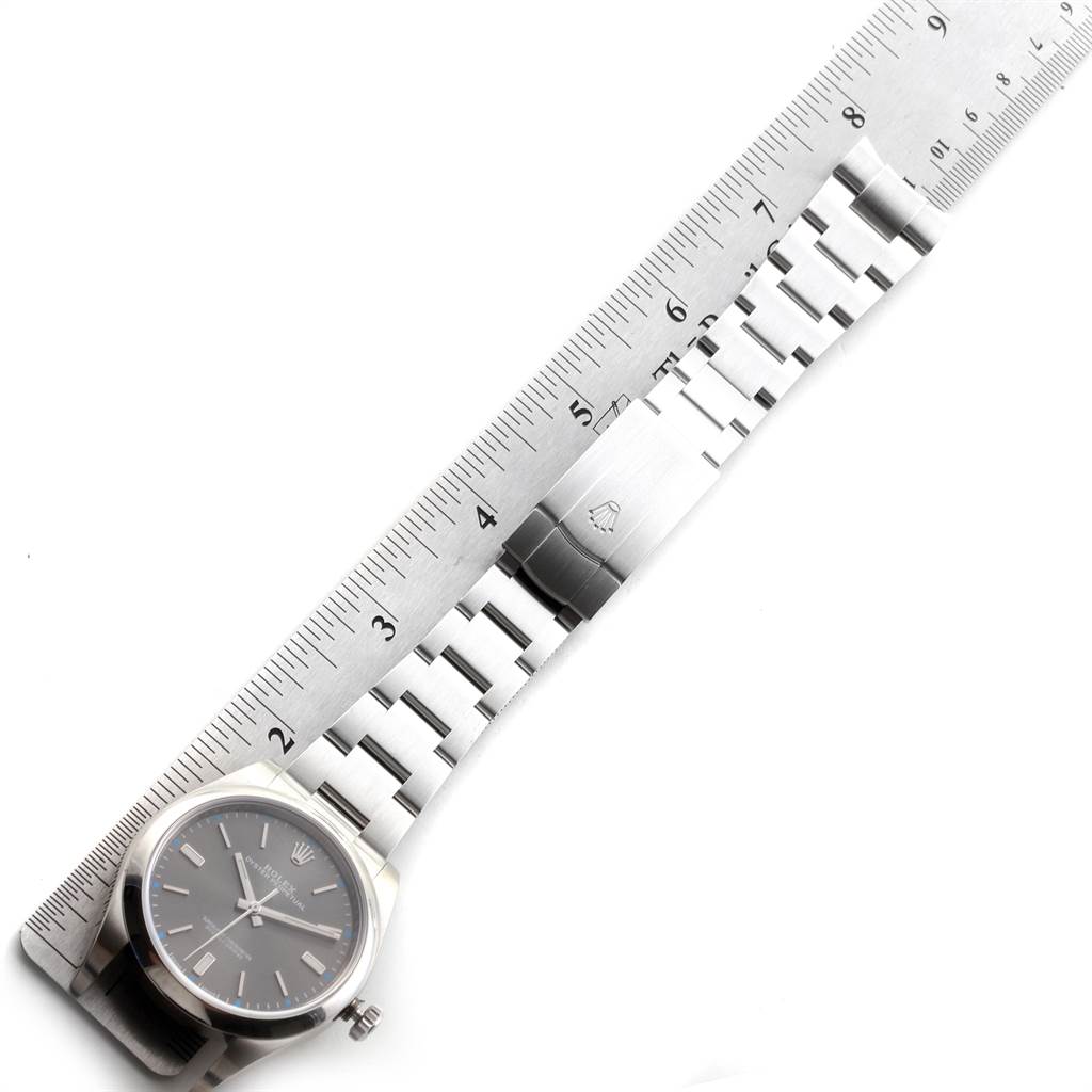 The image shows a Rolex Oyster Perpetual 114300 Men
s Stainless Steel Grey Dial 114300 Men
s Stainless Steel Grey Dial watch and its metal bracelet laid flat next to a ruler.