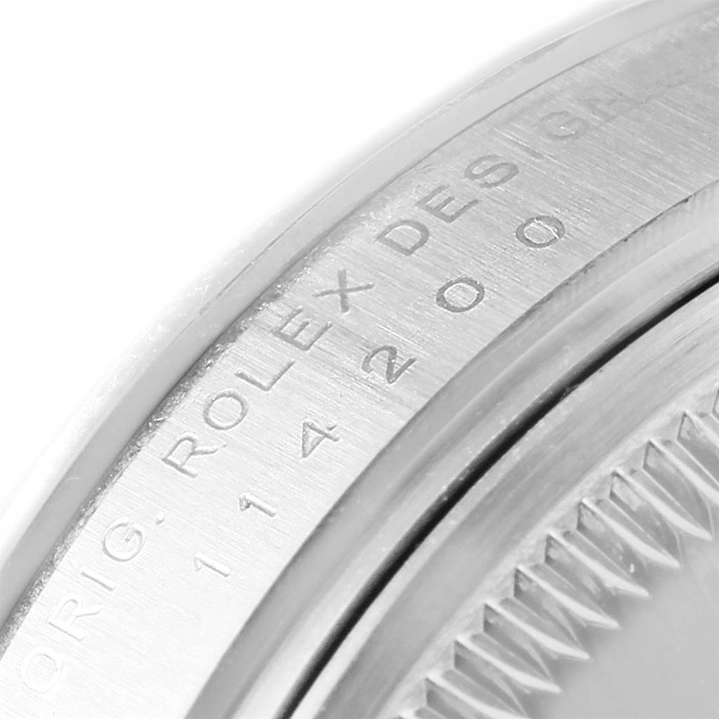 The image shows a close-up of the case back edge of a Rolex Oyster Perpetual 114200 Men
s Stainless Steel Black Dial 114200 Men
s Stainless Steel Black Dial watch with engraved serial numbers and text.