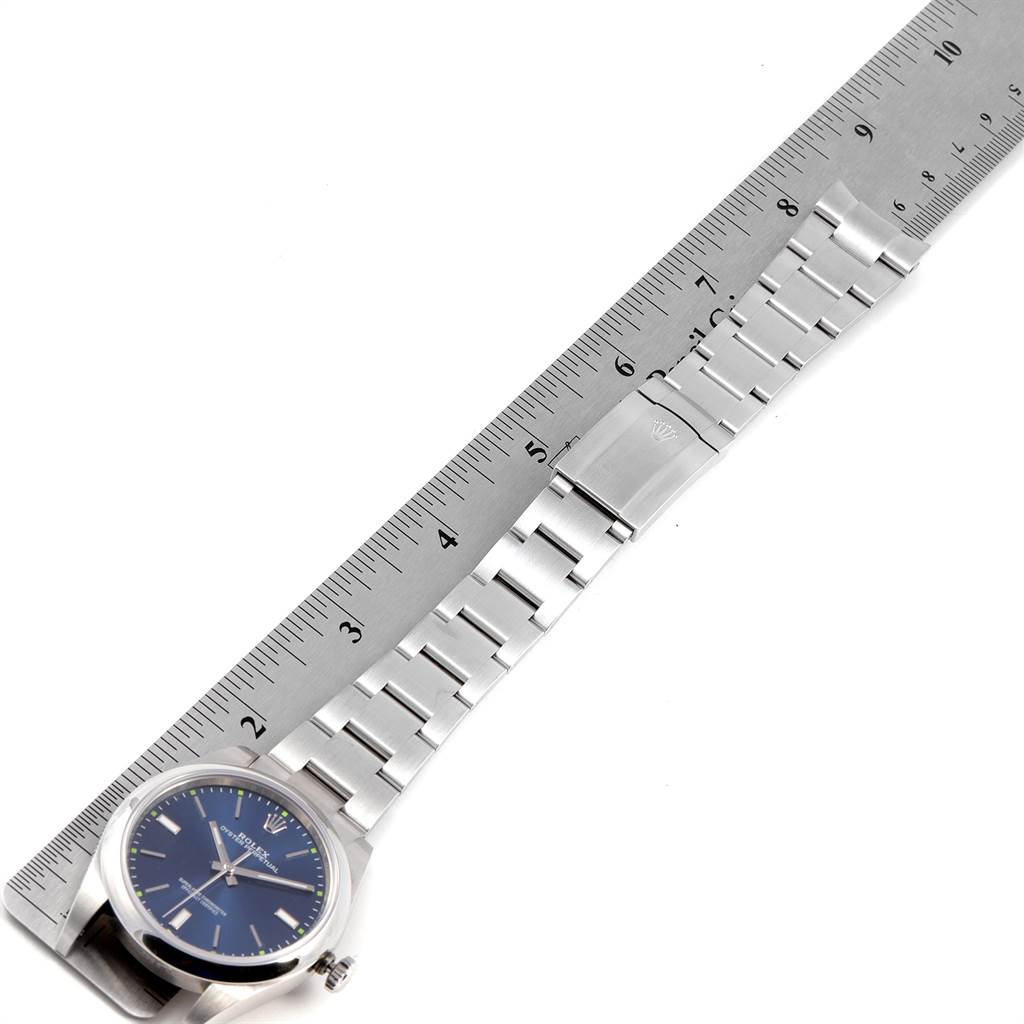 This image shows a top-down view of the Rolex Oyster Perpetual 114300 Men's Stainless Steel Blue Dial 114300 Men's Stainless Steel Blue Dial watch, displaying its blue dial, metal bracelet, and clasp alongside a ruler.