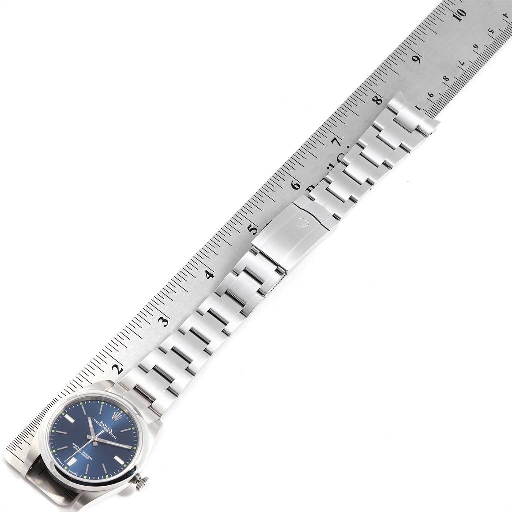 The image shows a side-angle view of a Rolex Oyster Perpetual 114300 Men's Stainless Steel Blue Dial 114300 Men's Stainless Steel Blue Dial watch with a blue dial and its stainless steel bracelet laid alongside a ruler.