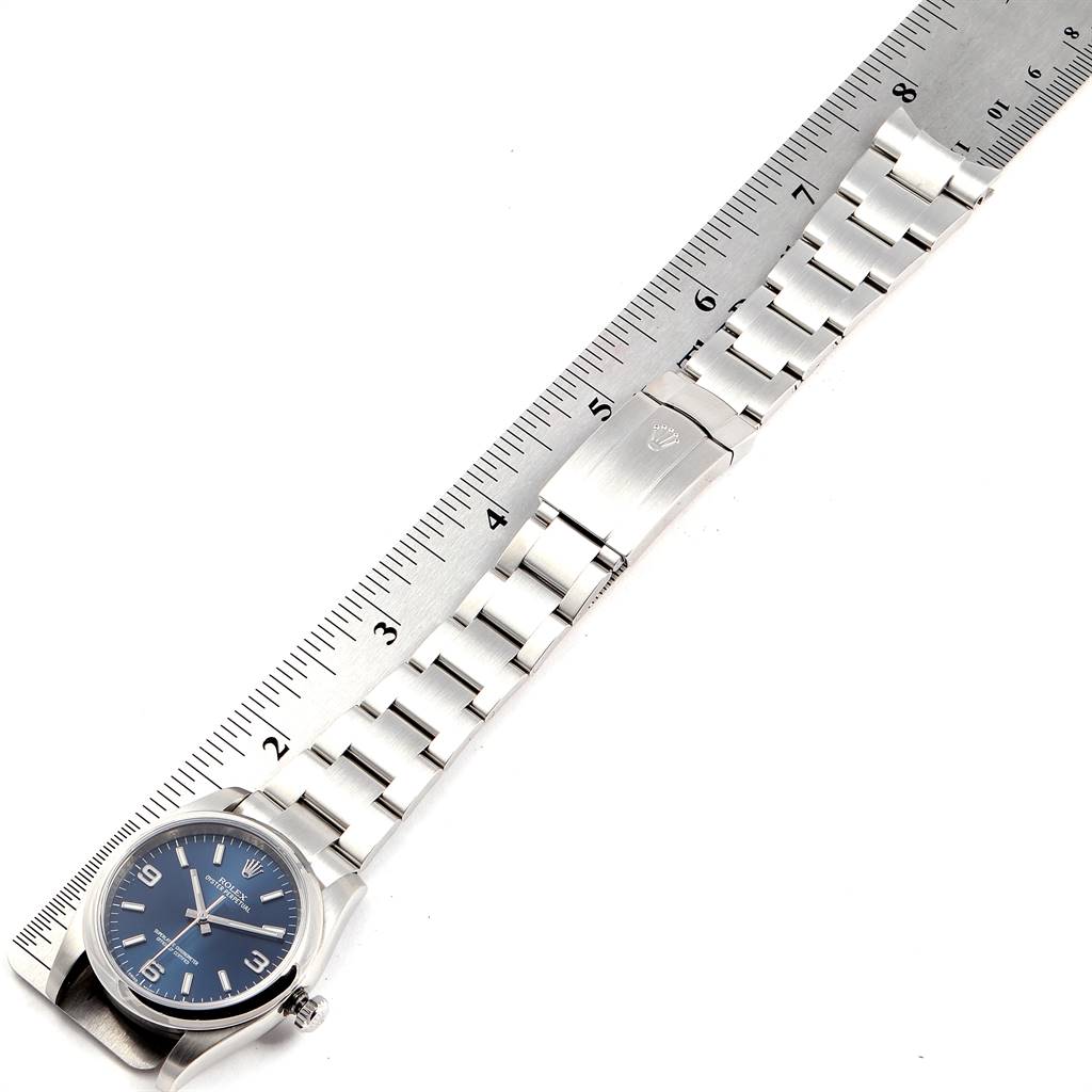The image shows a top view of a Rolex Oyster Perpetual 116000 Men
s Stainless Steel Blue Dial 116000 Men
s Stainless Steel Blue Dial watch with a blue dial and its stainless steel bracelet placed next to a ruler.