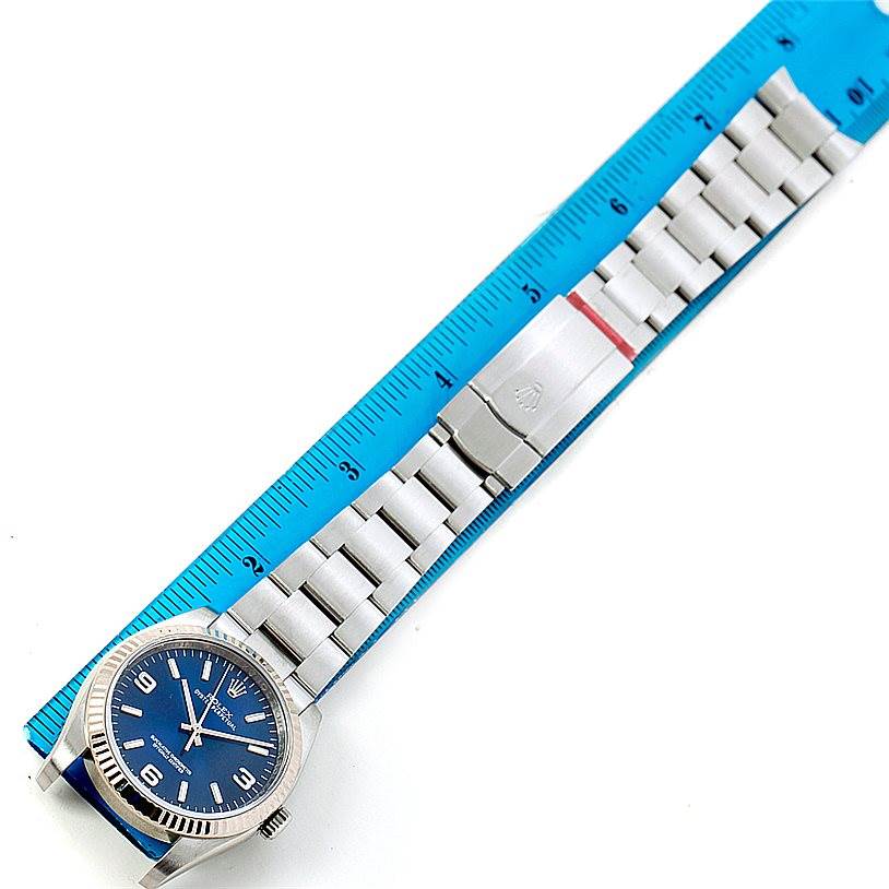 The image shows a top-down view of a Rolex Oyster Perpetual 116034 Men's Stainless Steel Blue Dial 116034 Men's Stainless Steel Blue Dial watch with a blue face and a stainless steel bracelet, next to a blue ruler.