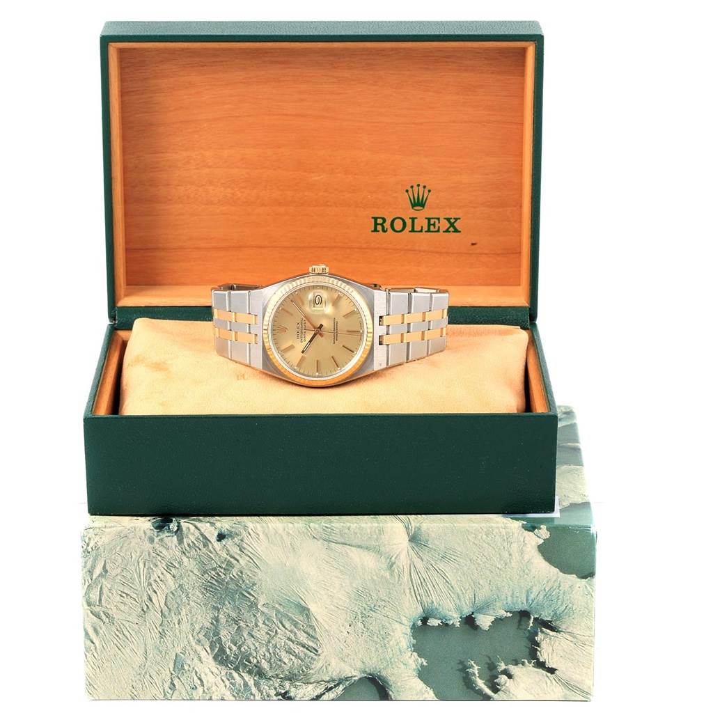 The Rolex Oysterquartz 17013 Men's Stainless Steel Champagne Dial 17013 Men's Stainless Steel Champagne Dial watch is pictured from a front angle inside its green and wooden branded box, atop an outer box with an abstract design.