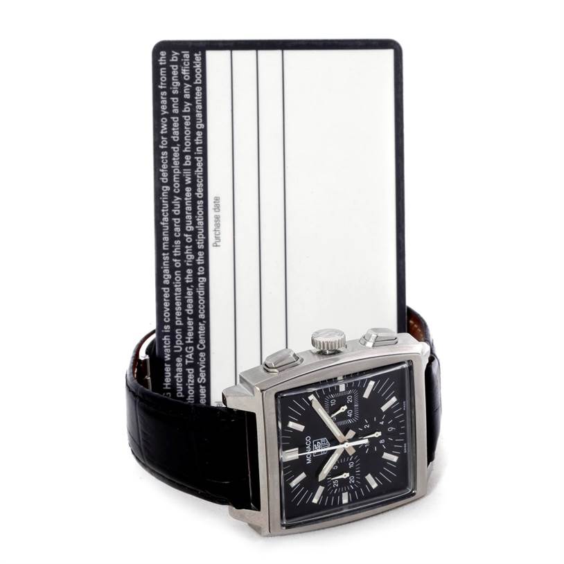 The image displays a Tag Heuer Monaco CW2111 Men
s Stainless Steel Black Dial CW2111 Men
s Stainless Steel Black Dial watch standing upright with the front face visible, against a warranty card in the background.