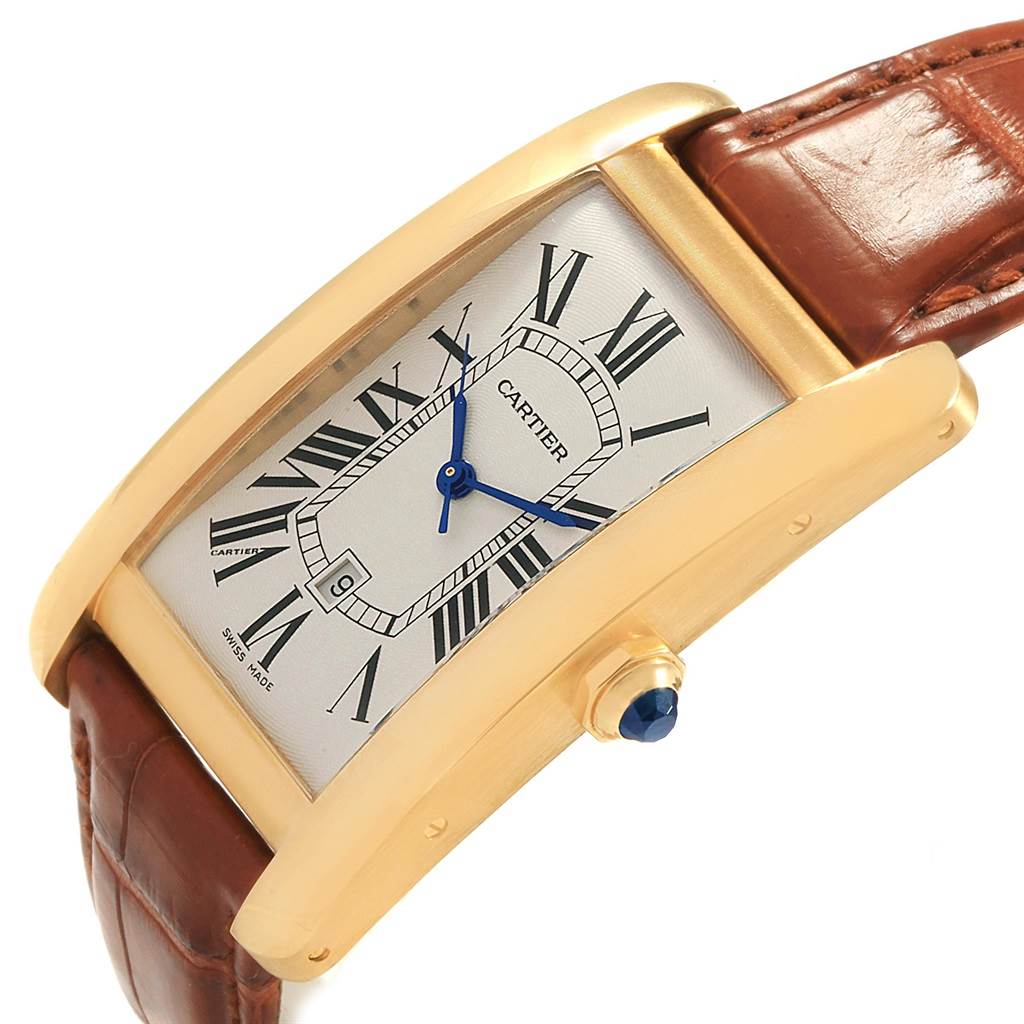 The image shows a Cartier Tank Americaine W2603156 Men's Yellow Gold Silver Dial W2603156 Men's Yellow Gold Silver Dial watch at an angle, displaying its dial, blue hands, Roman numerals, gold case, and brown leather strap.